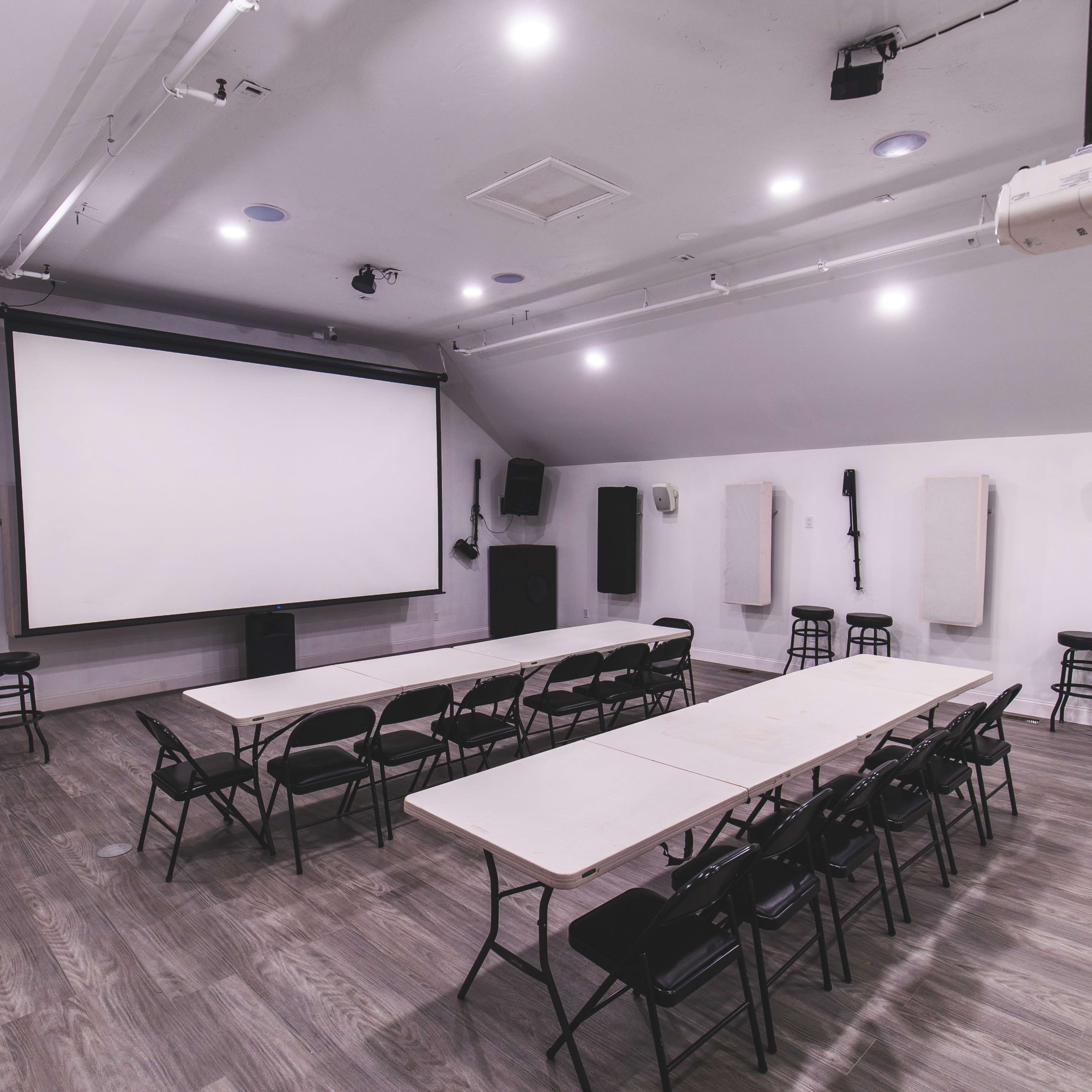 An empty multi-purpose room with a large screen at the front, sound equipment on the sides, and rows of chairs arranged around several tables.