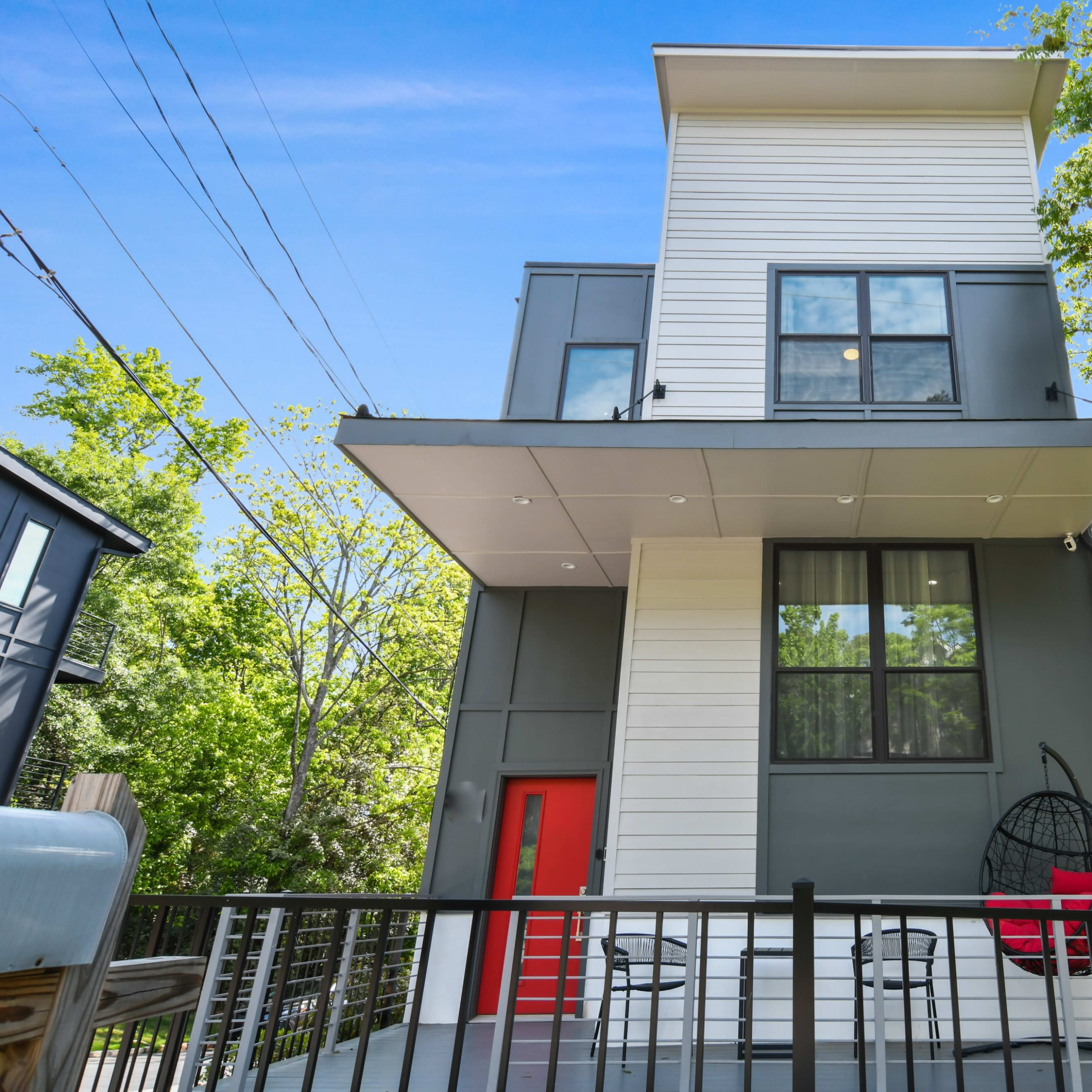 A modern two-story house with a flat roof, featuring a red front door and a balcony, surrounded by trees.