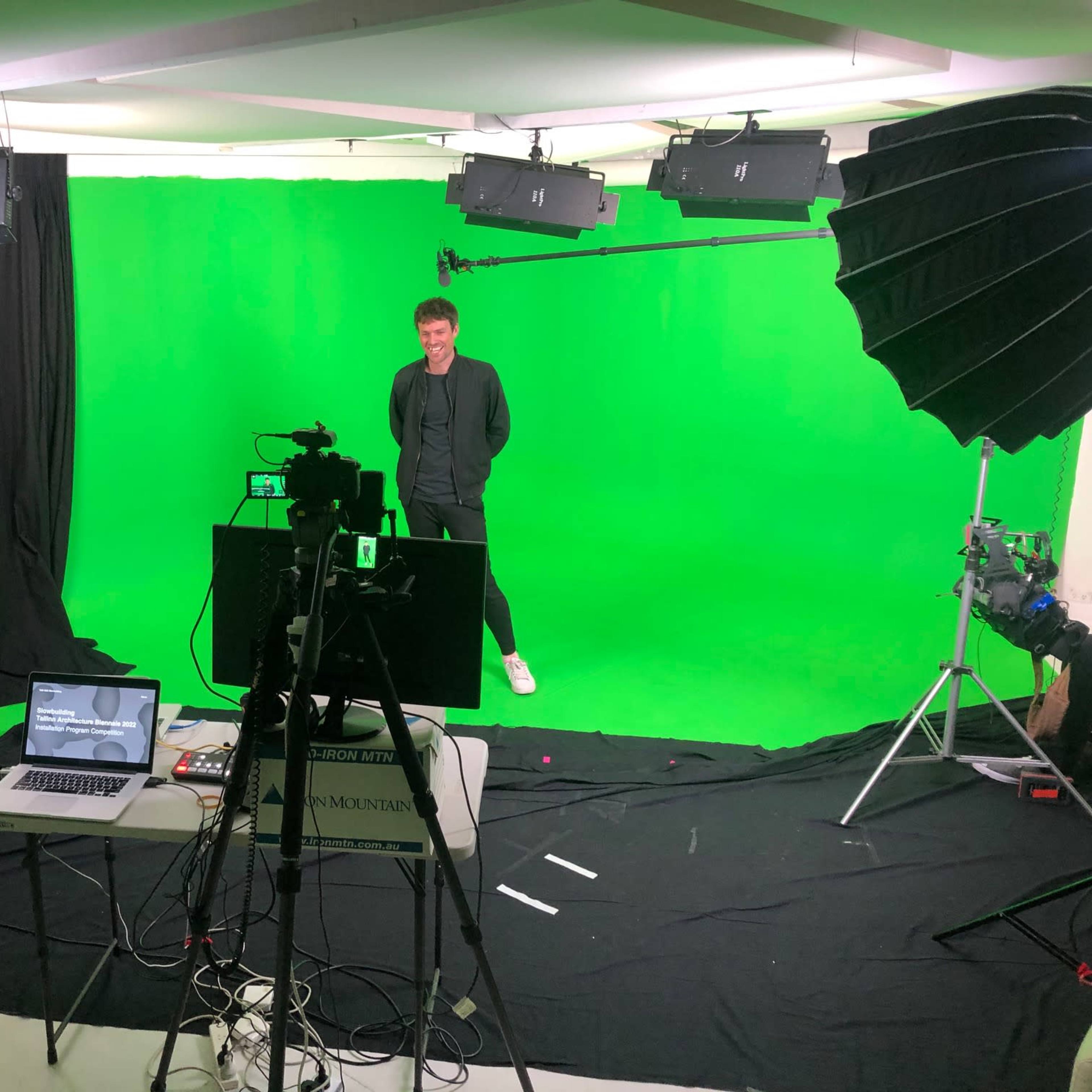 A person stands on a green screen set, surrounded by studio lights and camera equipment, with a laptop placed on a table in front.