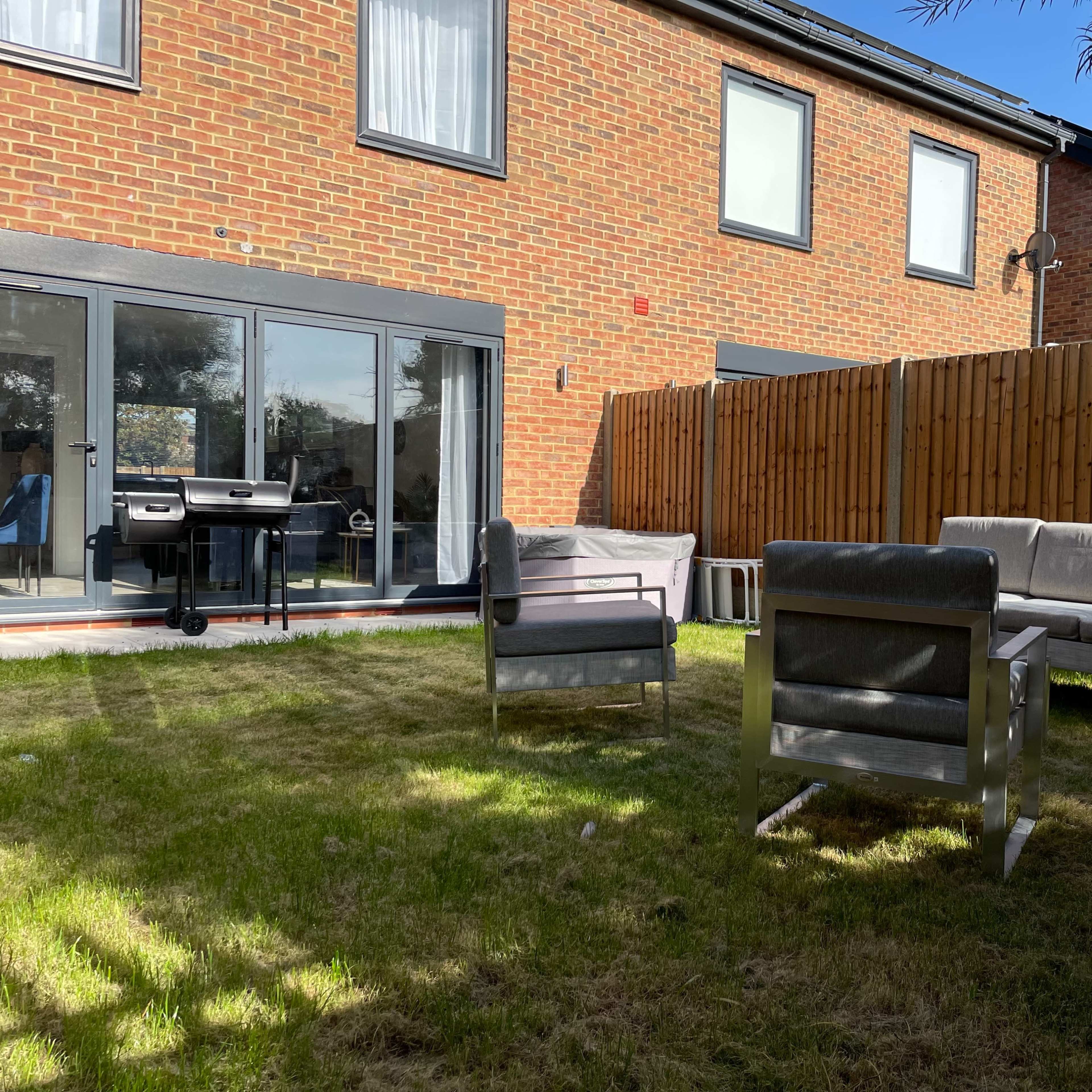 A small backyard with a grassy area, featuring four outdoor seating chairs arranged around a central space and a barbecue grill near a glass door leading into a brick building.