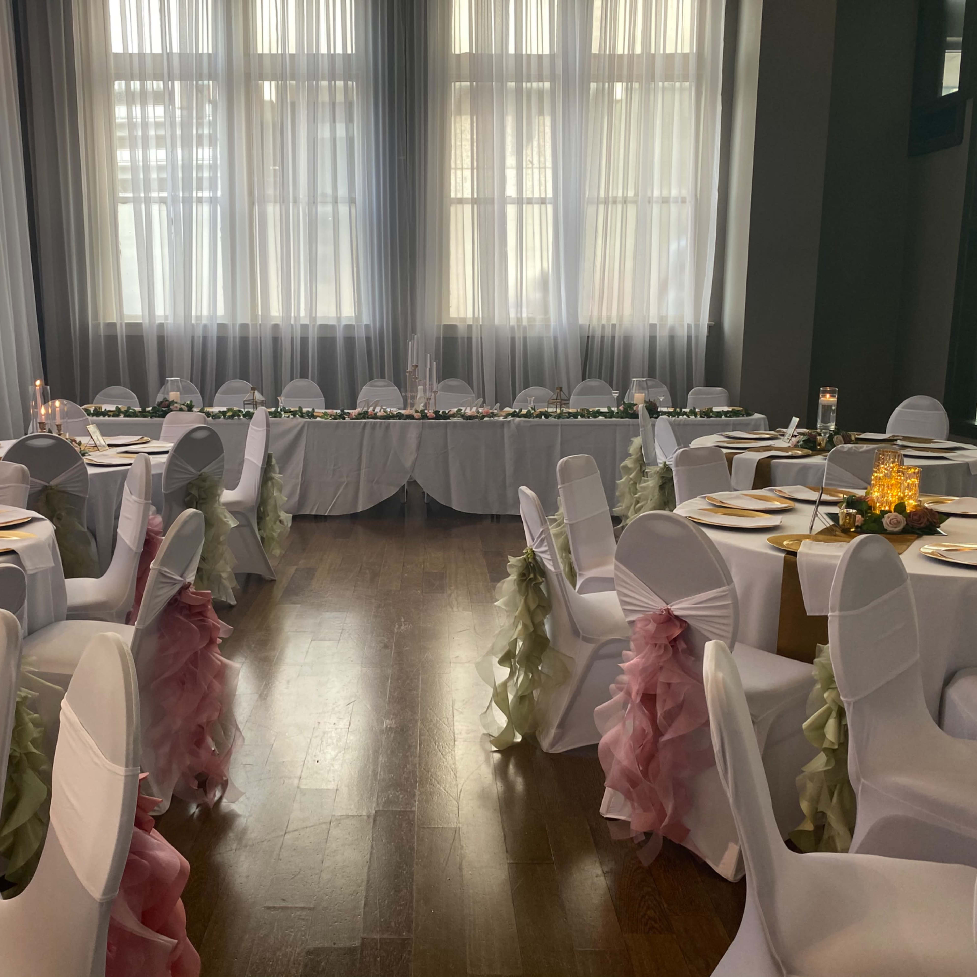 A banquet hall set up for an event, featuring tables adorned with white tablecloths, plates, and chairs draped in fabric with pink and green sashes.