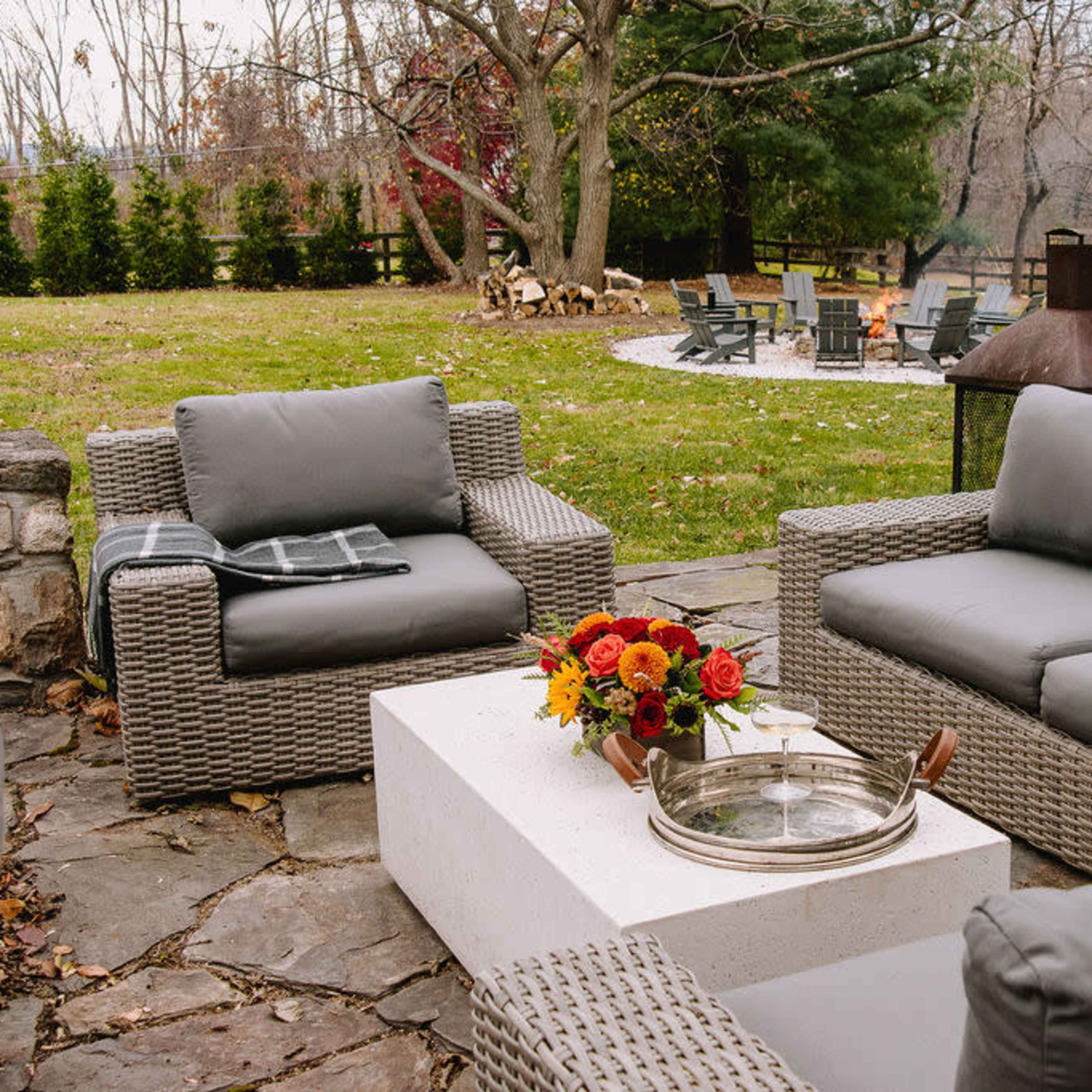 A seating area with gray wicker furniture surrounds a stone table featuring a tray, with a fire pit and grassy landscape visible in the background.
