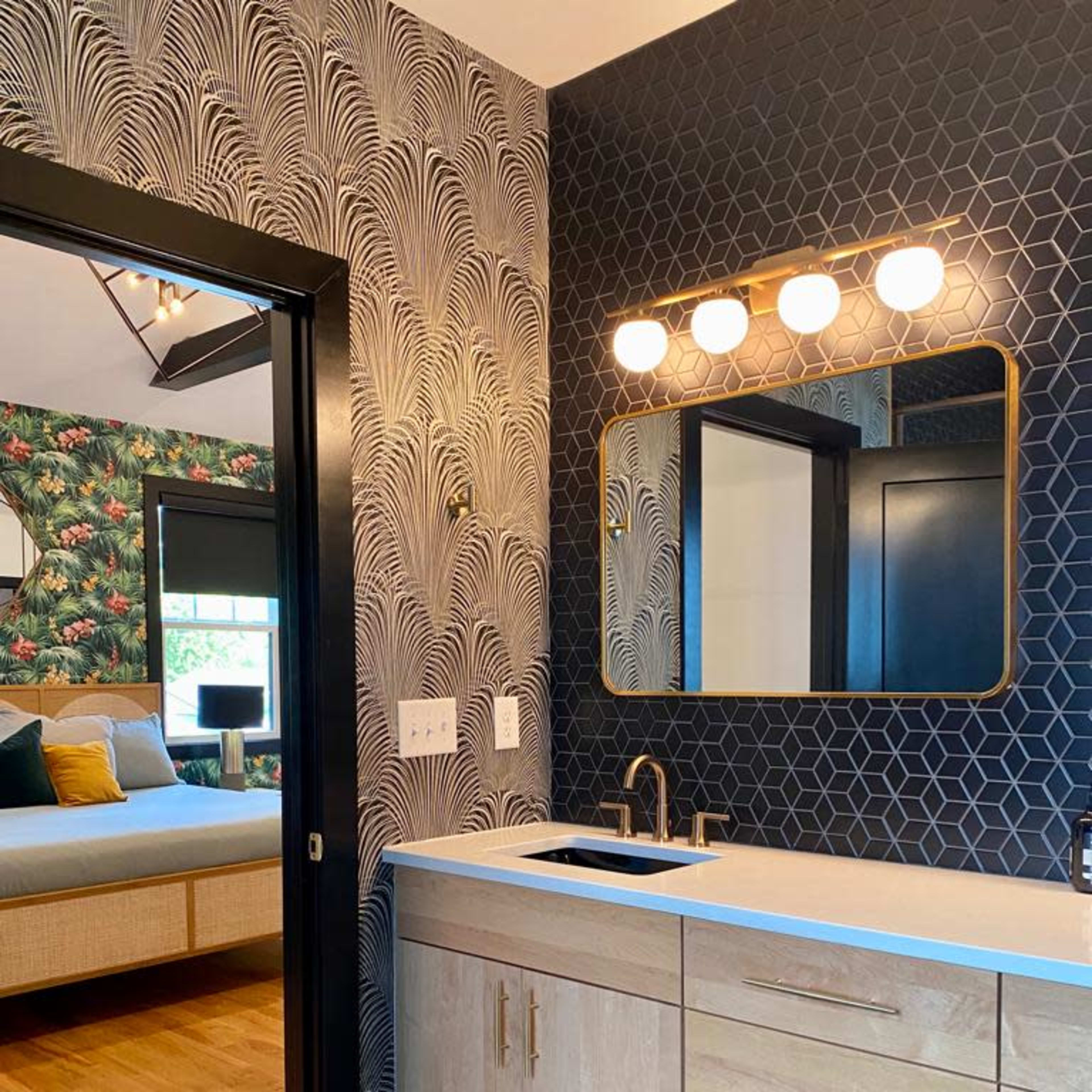 A modern bathroom featuring a vanity with a white countertop, wooden cabinets, a round mirror, and a geometric tile floor, adjacent to a bedroom with floral wallpaper.