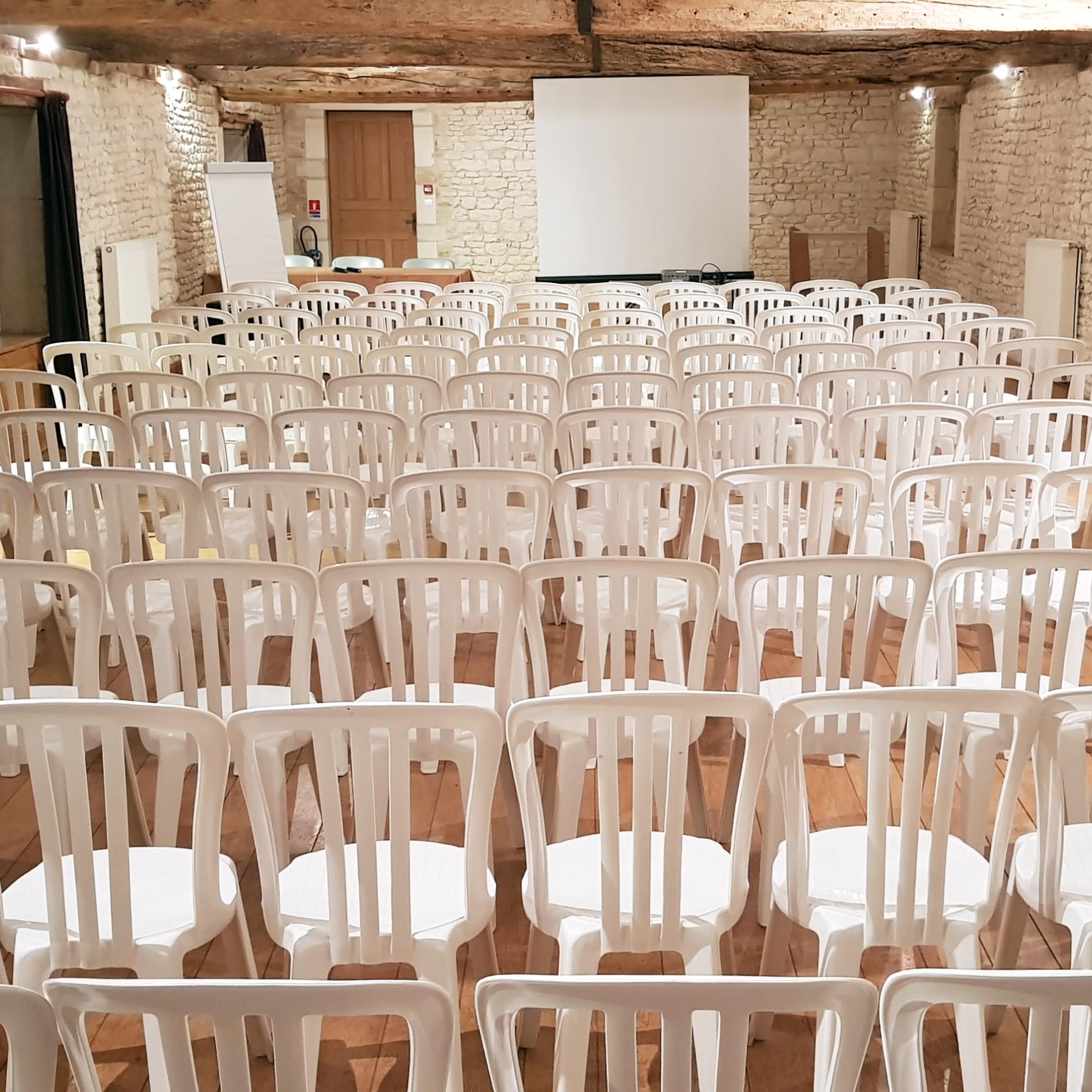 A room with stone walls and wooden beams contains rows of white plastic chairs arranged for an event.