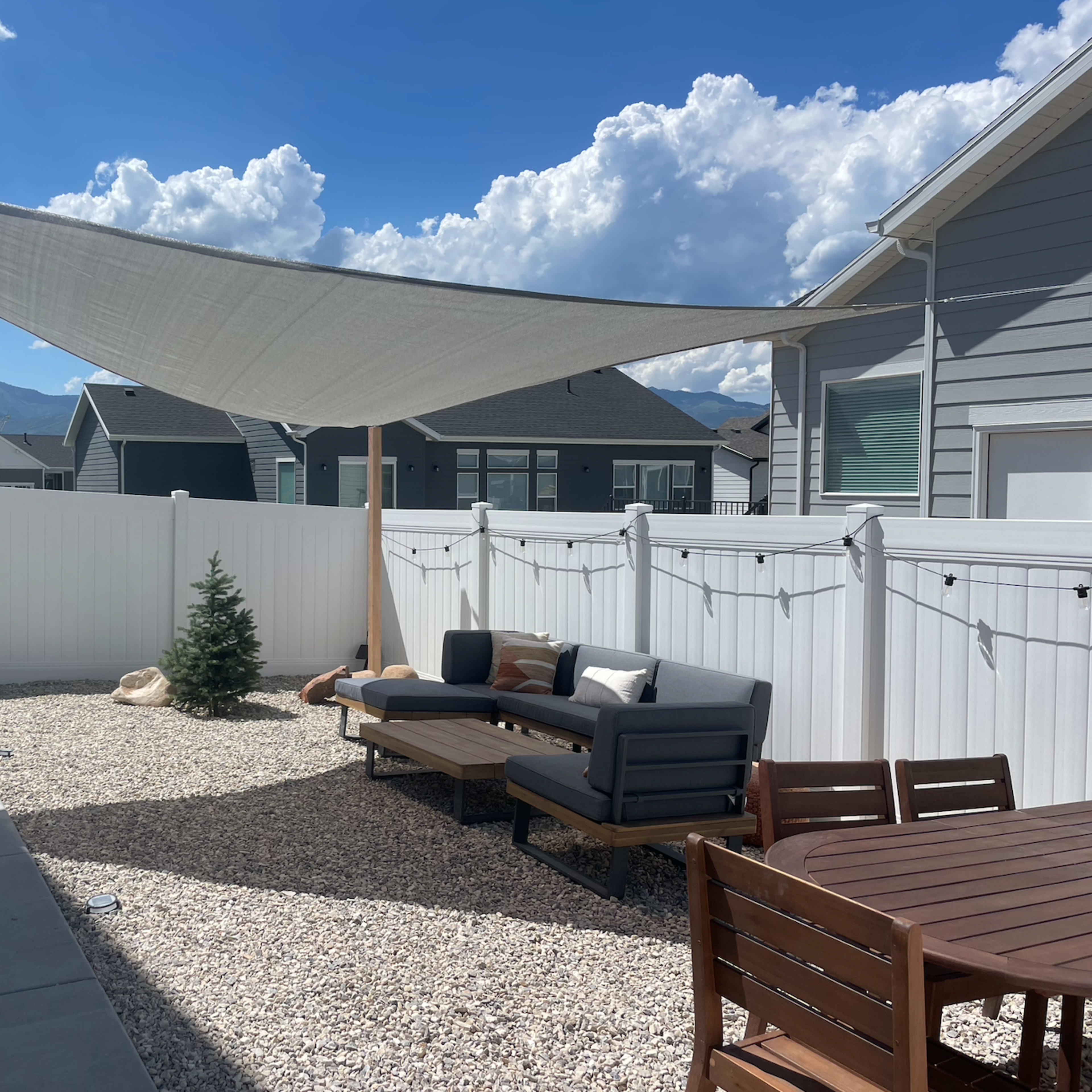 A gravel backyard with a shaded seating area, a small tree, and string lights, bordered by a white fence and houses in the background.