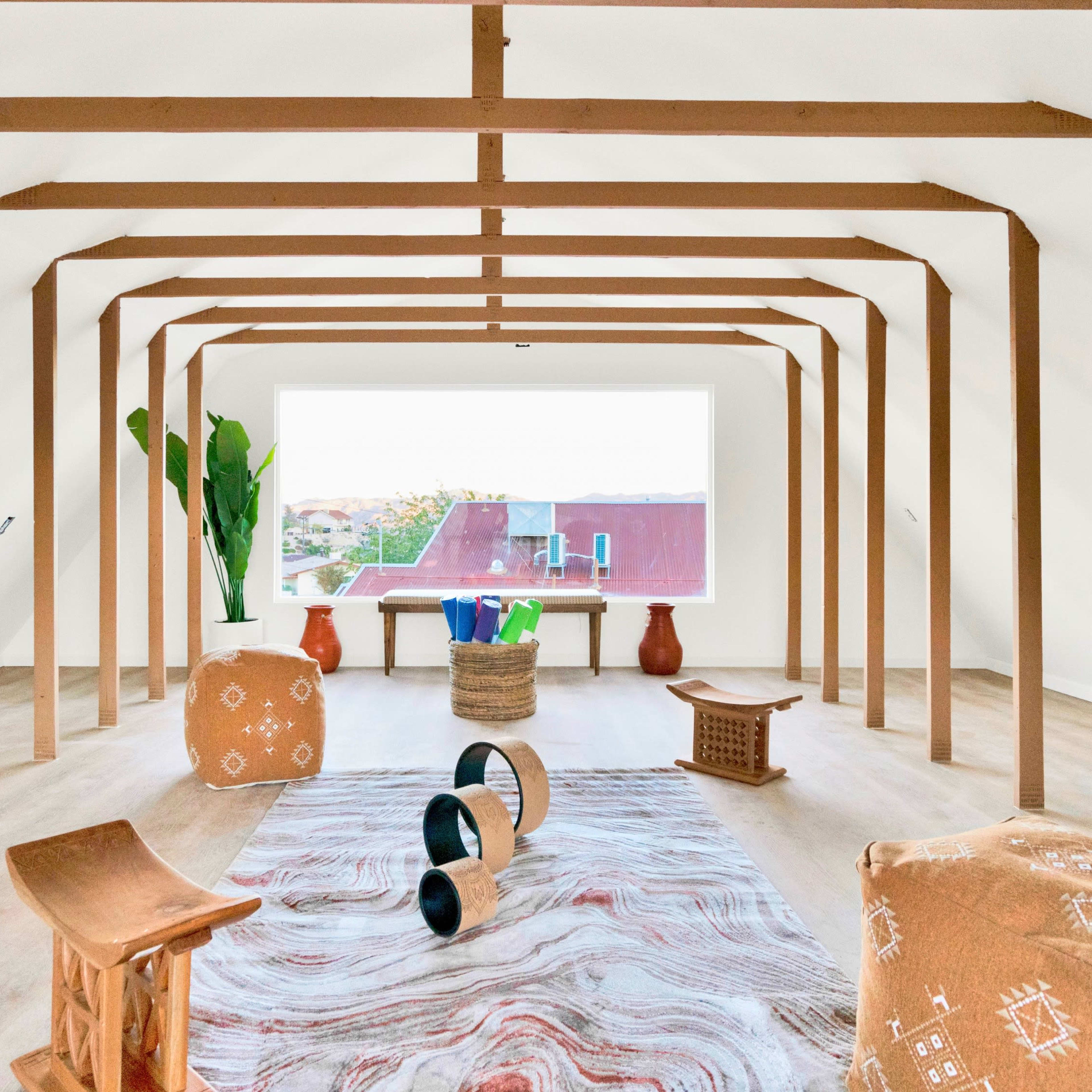 A spacious, minimalistic room with wooden beams, various furniture pieces, a large window, and plants.