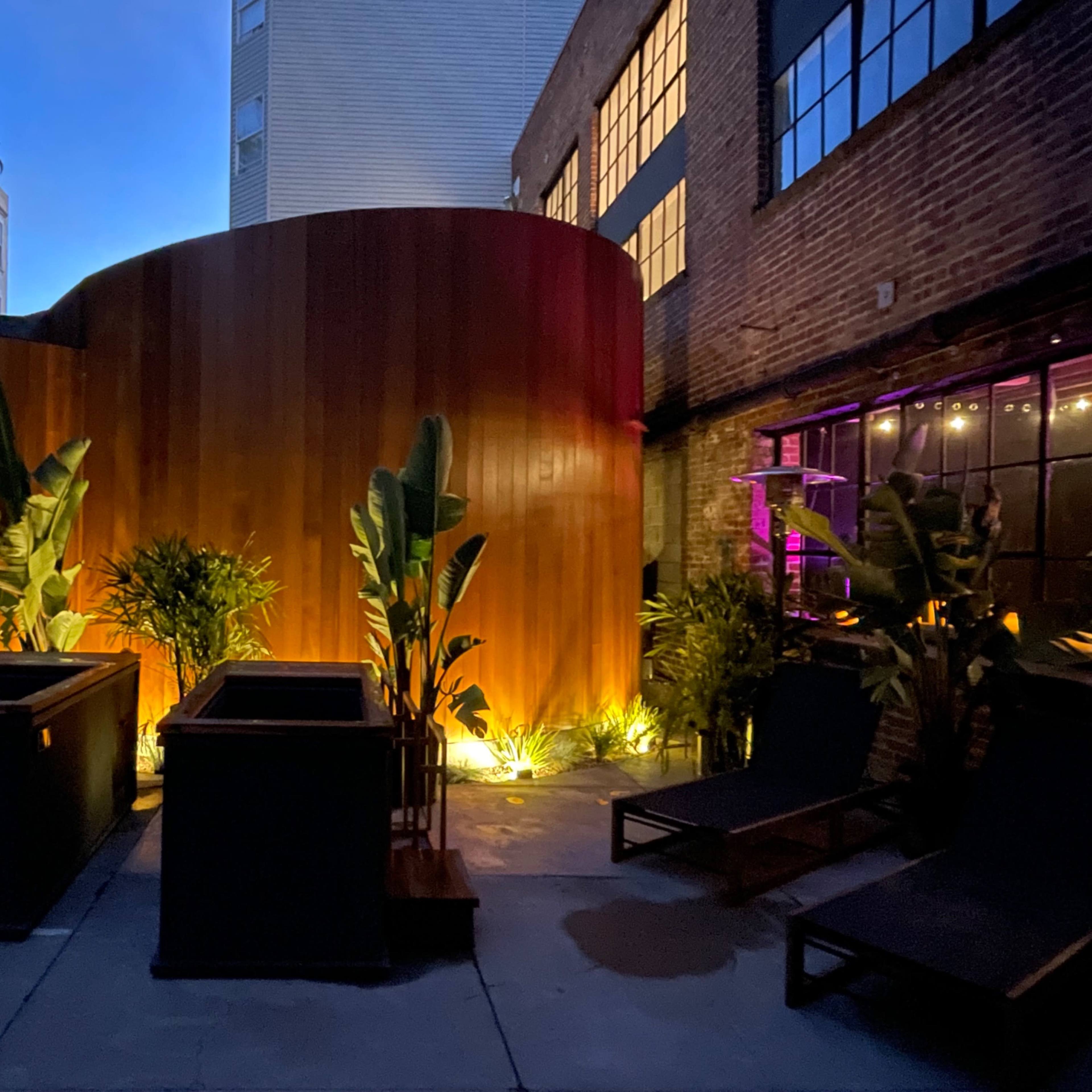 A dimly lit outdoor space with wooden walls, tropical plants, and sleek lounge chairs against a backdrop of a building with large windows.
