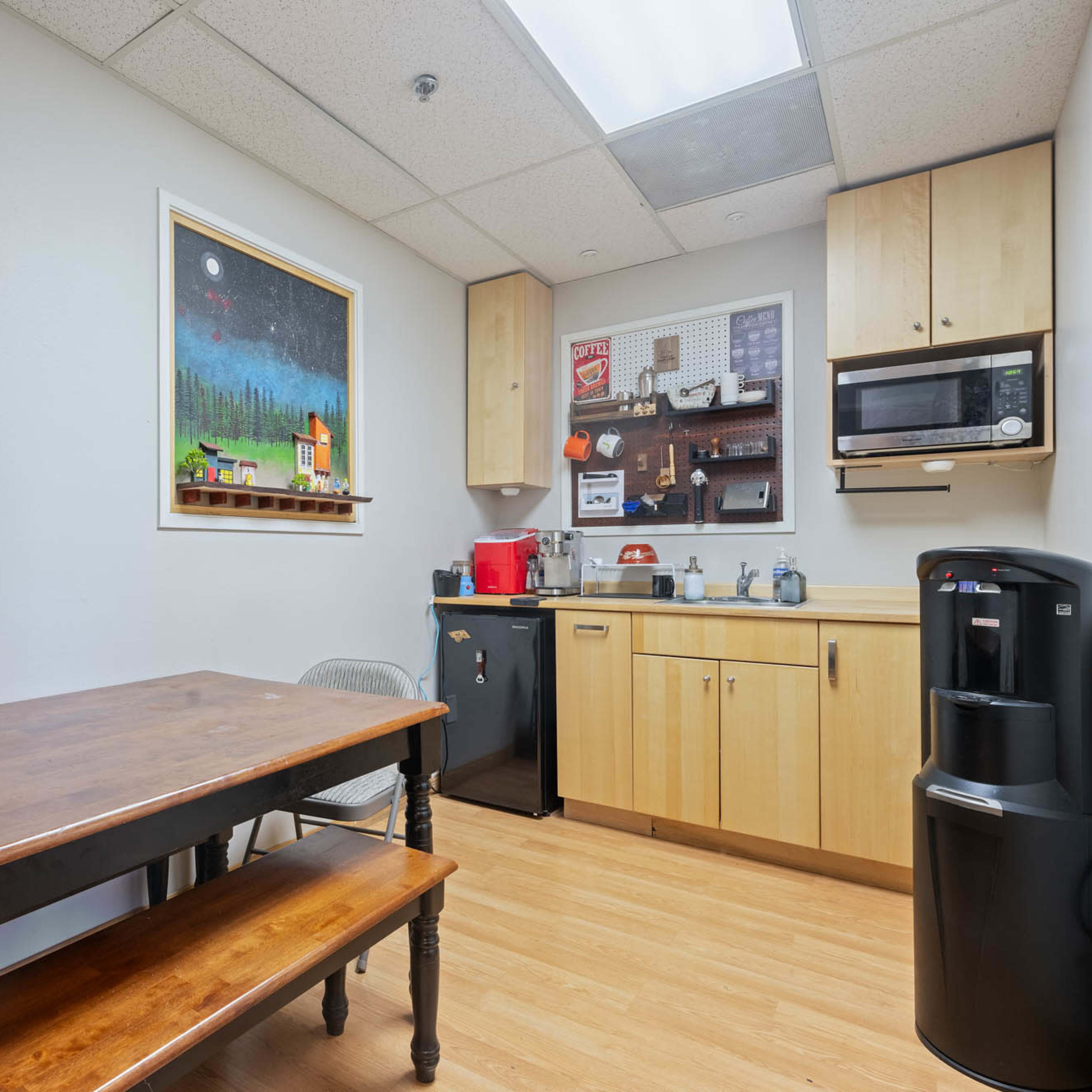 A small breakroom with a wooden table, chairs, and a kitchenette that includes a microwave, sink, and refrigerator.