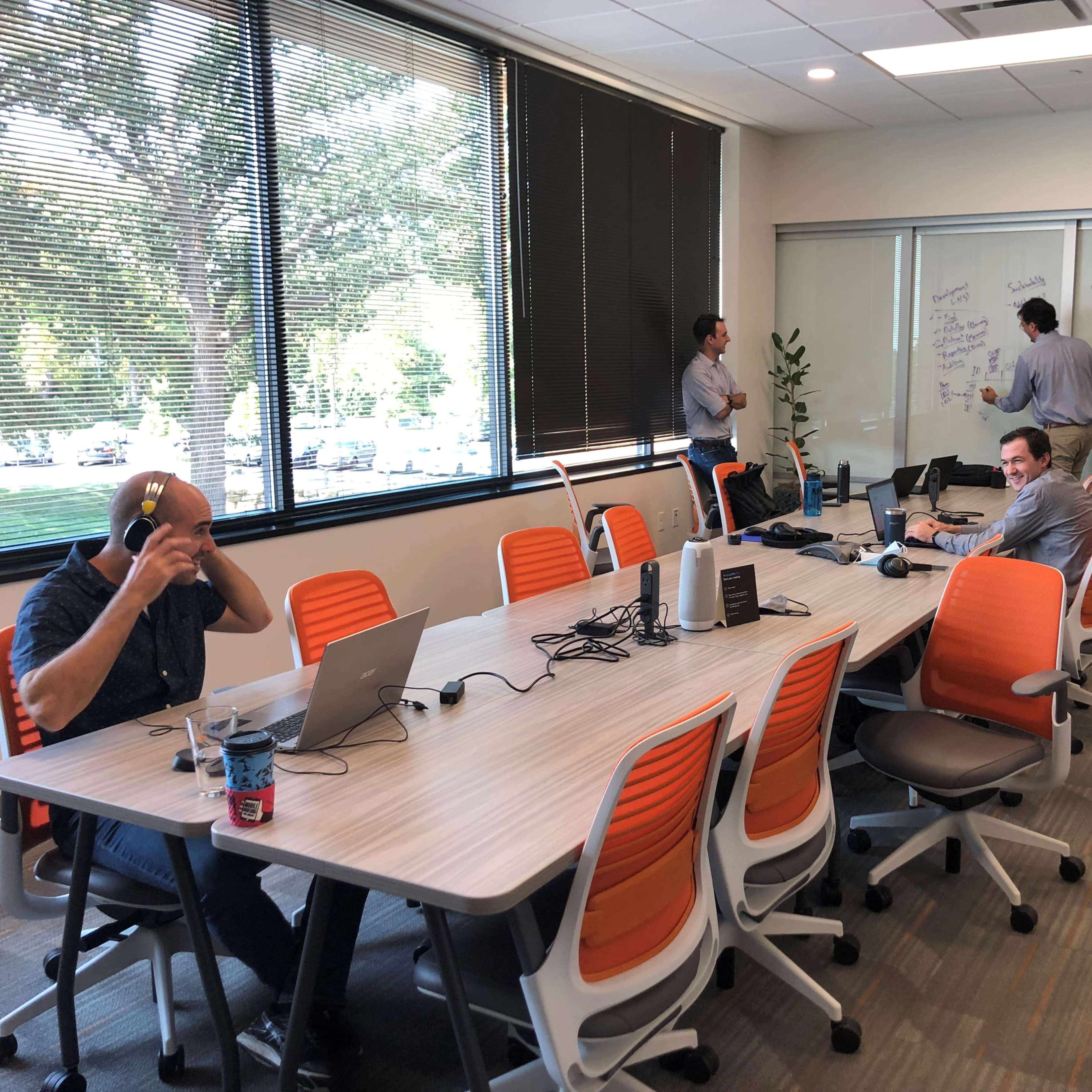Four people are working in a conference room, with two seated at a table using laptops and two standing near a whiteboard.