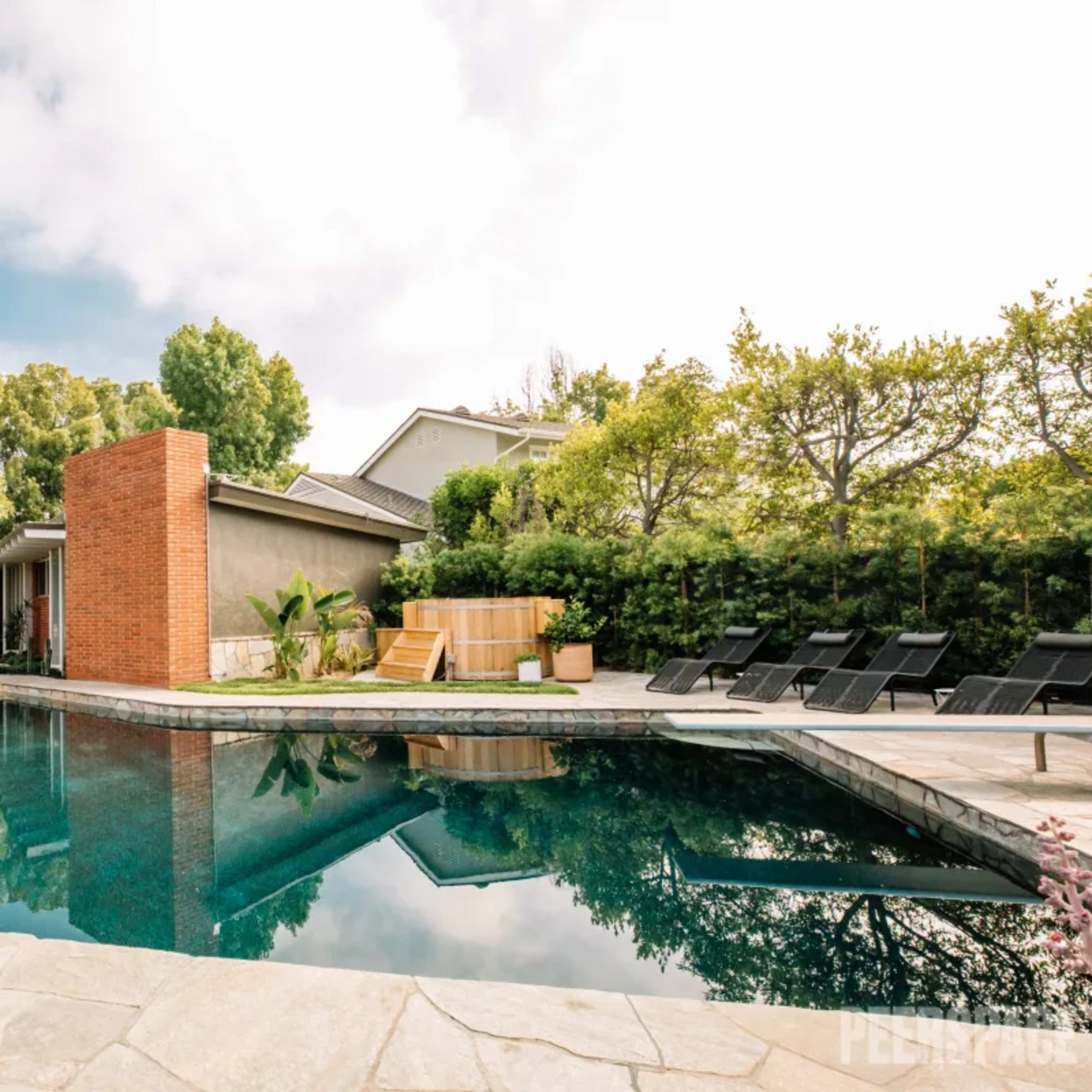 A modern outdoor pool surrounded by a stone patio, with a brick feature wall and lounge chairs nearby, set among lush greenery.