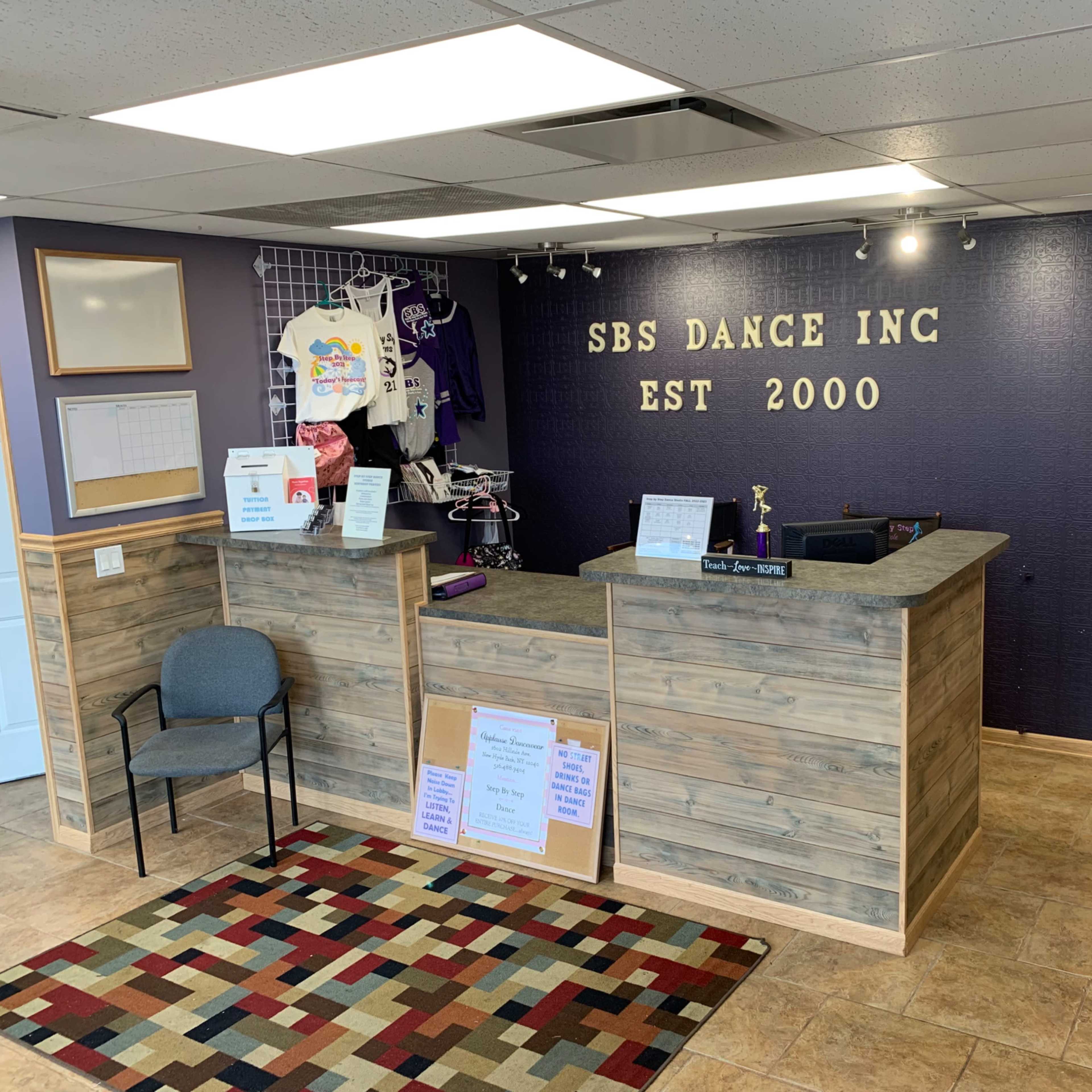 A reception area for SBS Dance Inc., featuring a wooden front desk, promotional items on display, and a seating area with a patterned rug.