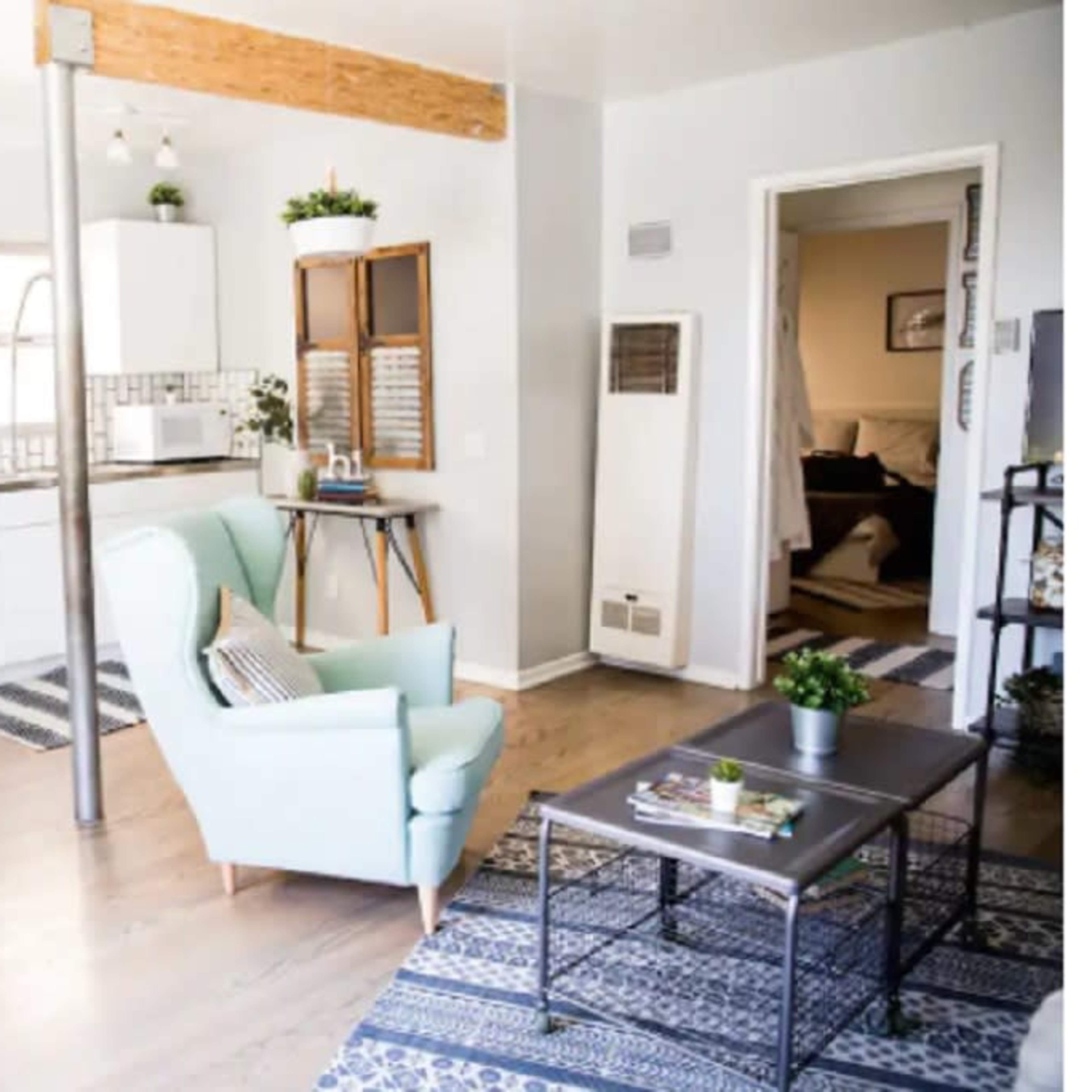 A modern kitchen and living area featuring a circular dining table with four stools, a green armchair, and a coffee table, all set against a backdrop of white cabinets and a patterned rug.
