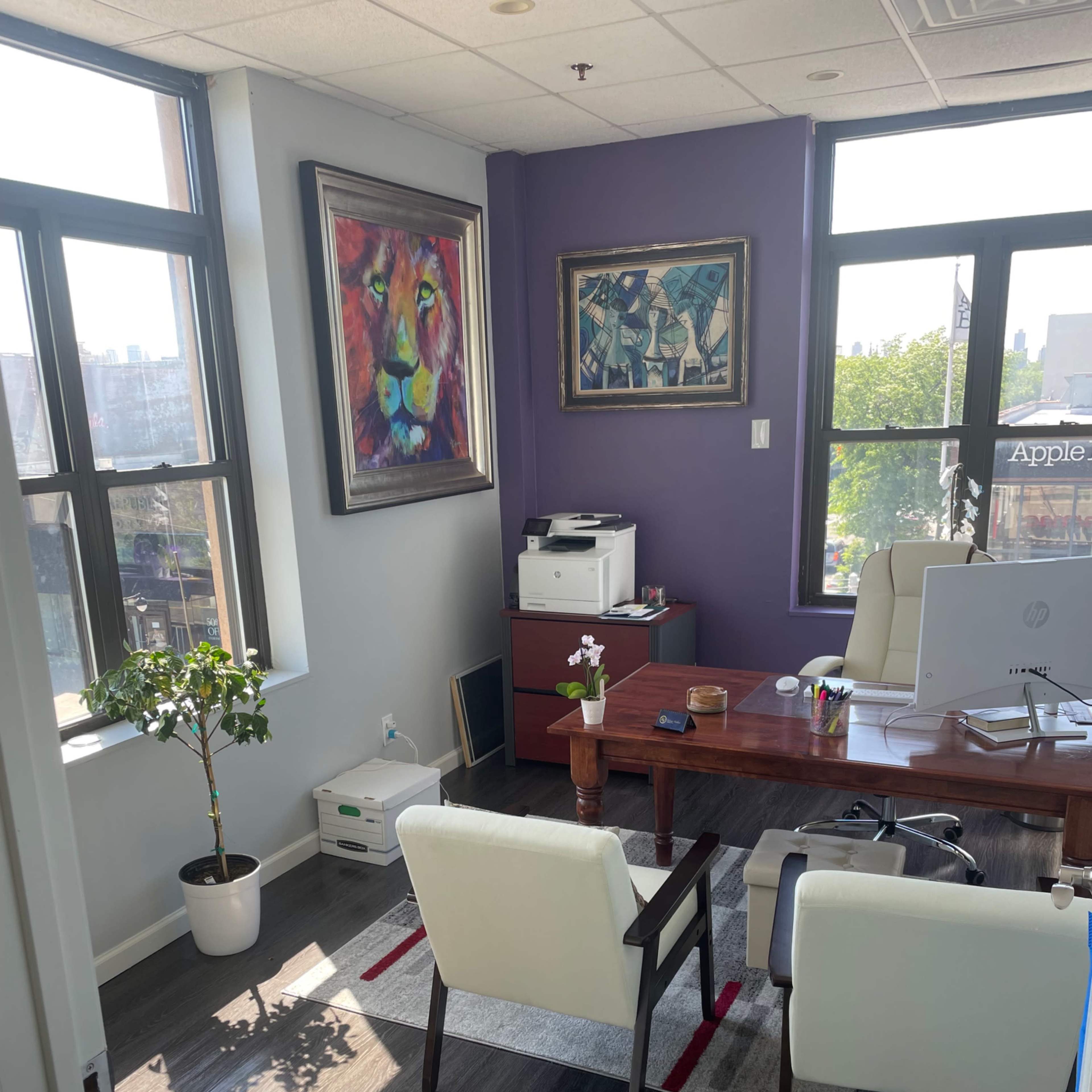 An office with a wooden desk, two white chairs, a potted plant, and artwork on the walls, featuring large windows that let in natural light.