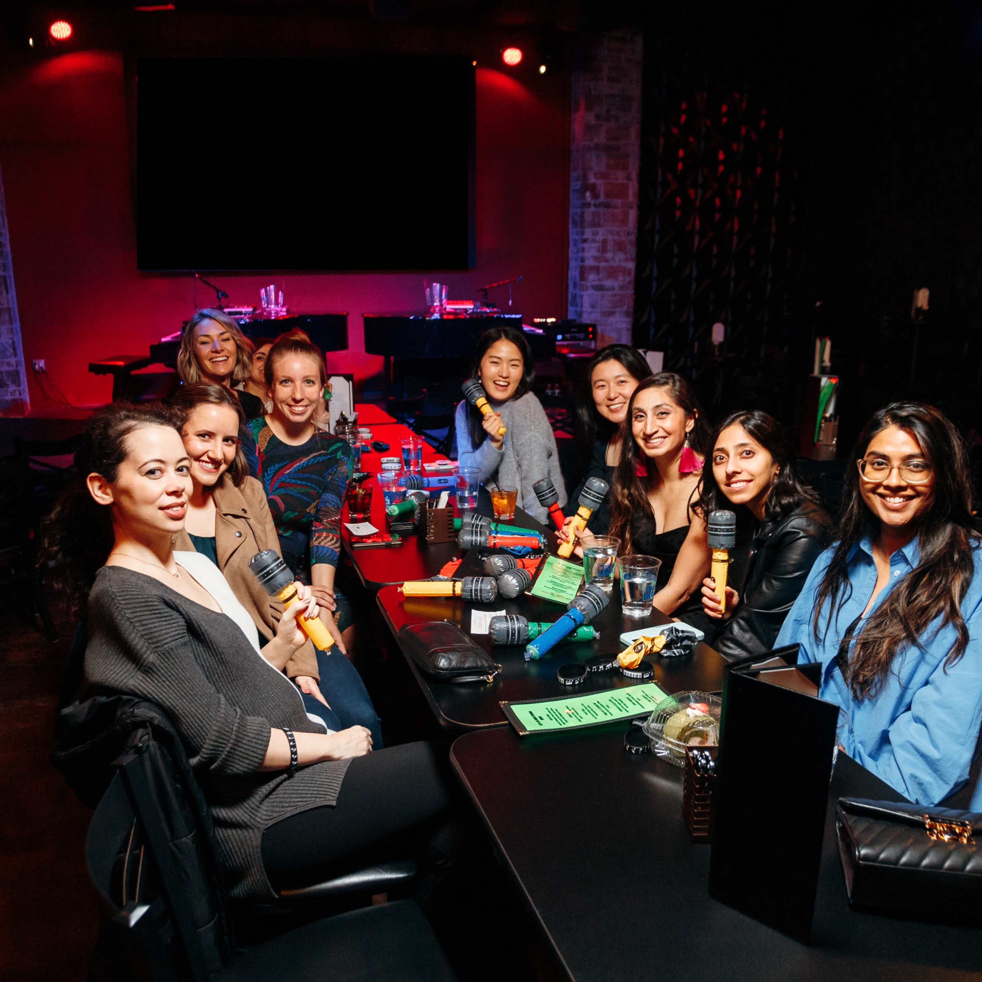 A group of nine women sits around a table in a dimly lit entertainment venue, each holding colorful microphones.