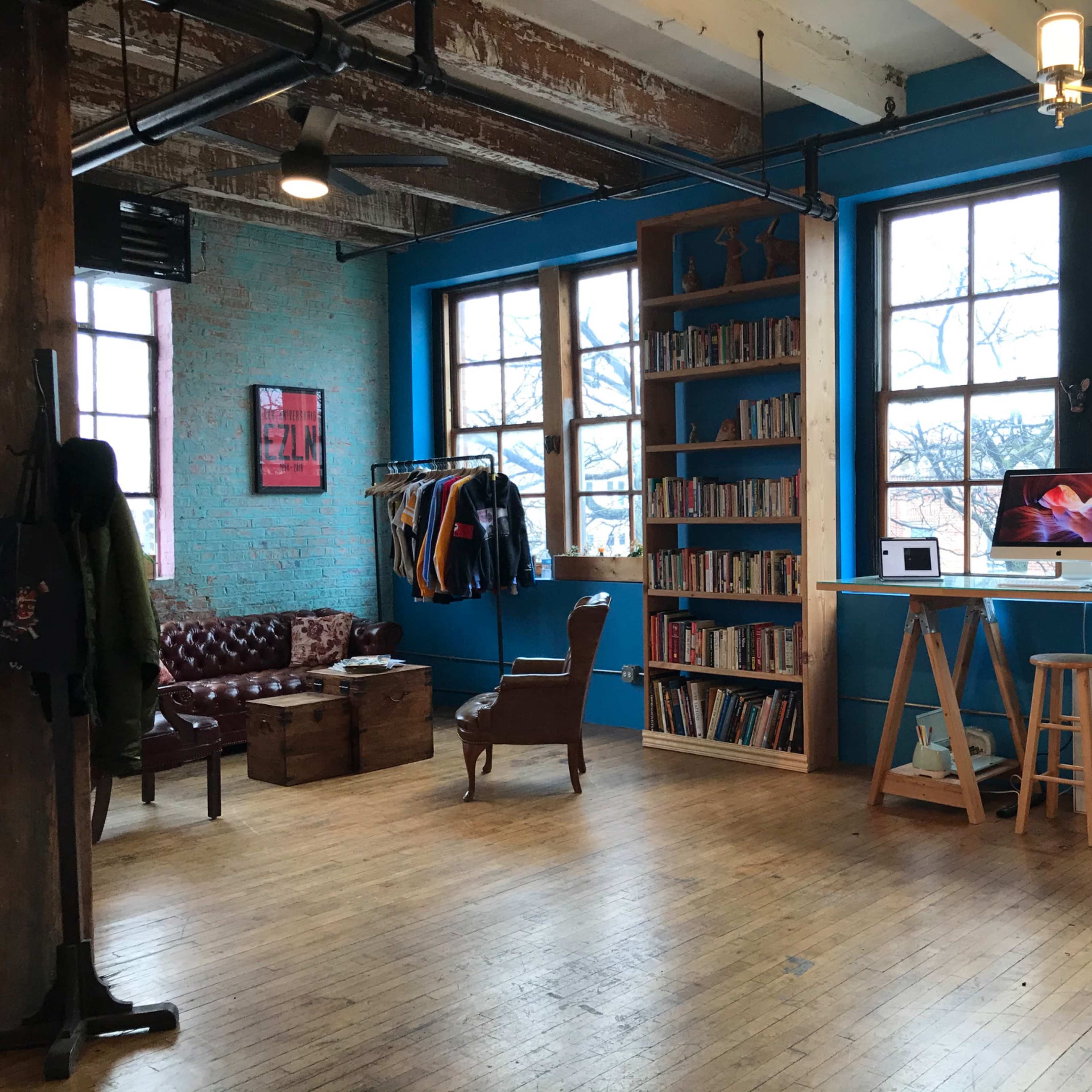 A spacious room with blue walls, wooden flooring, large windows, a bookshelf filled with books, a vintage armchair, and a computer setup on a desk.