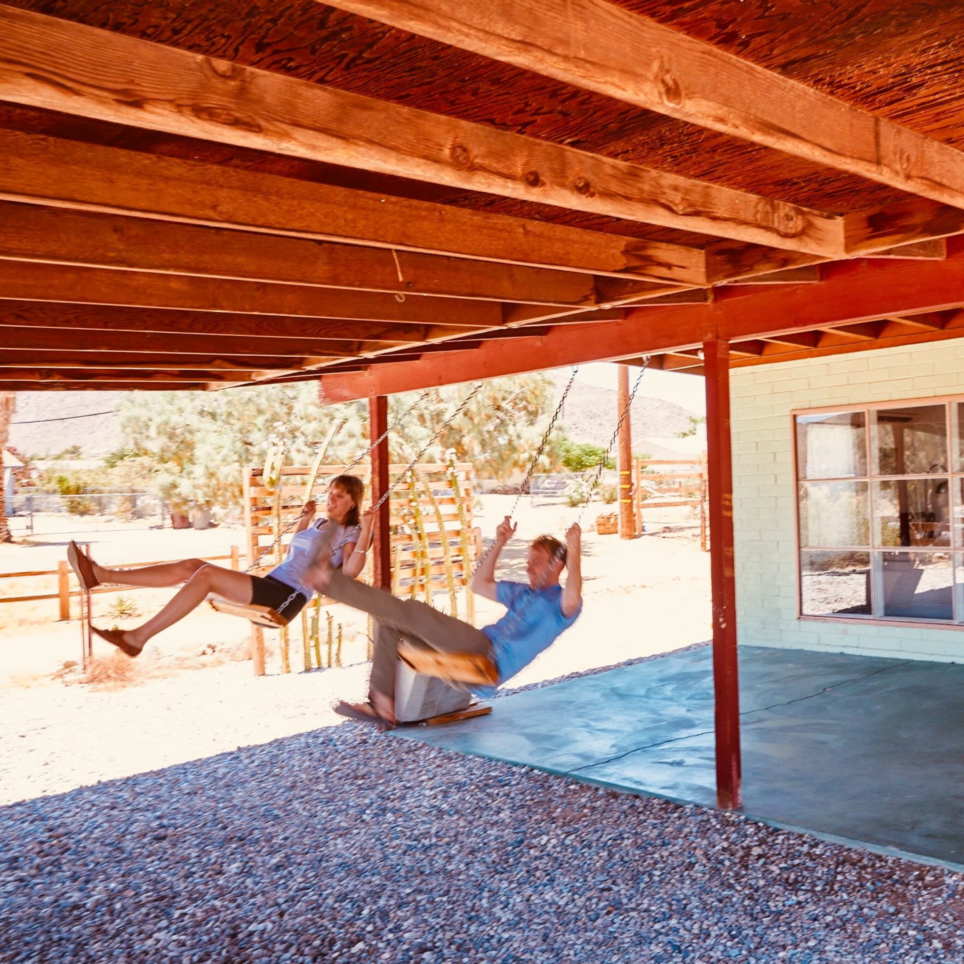 Two children are swinging on a rope swing beneath a wooden porch, with a desert landscape visible in the background.