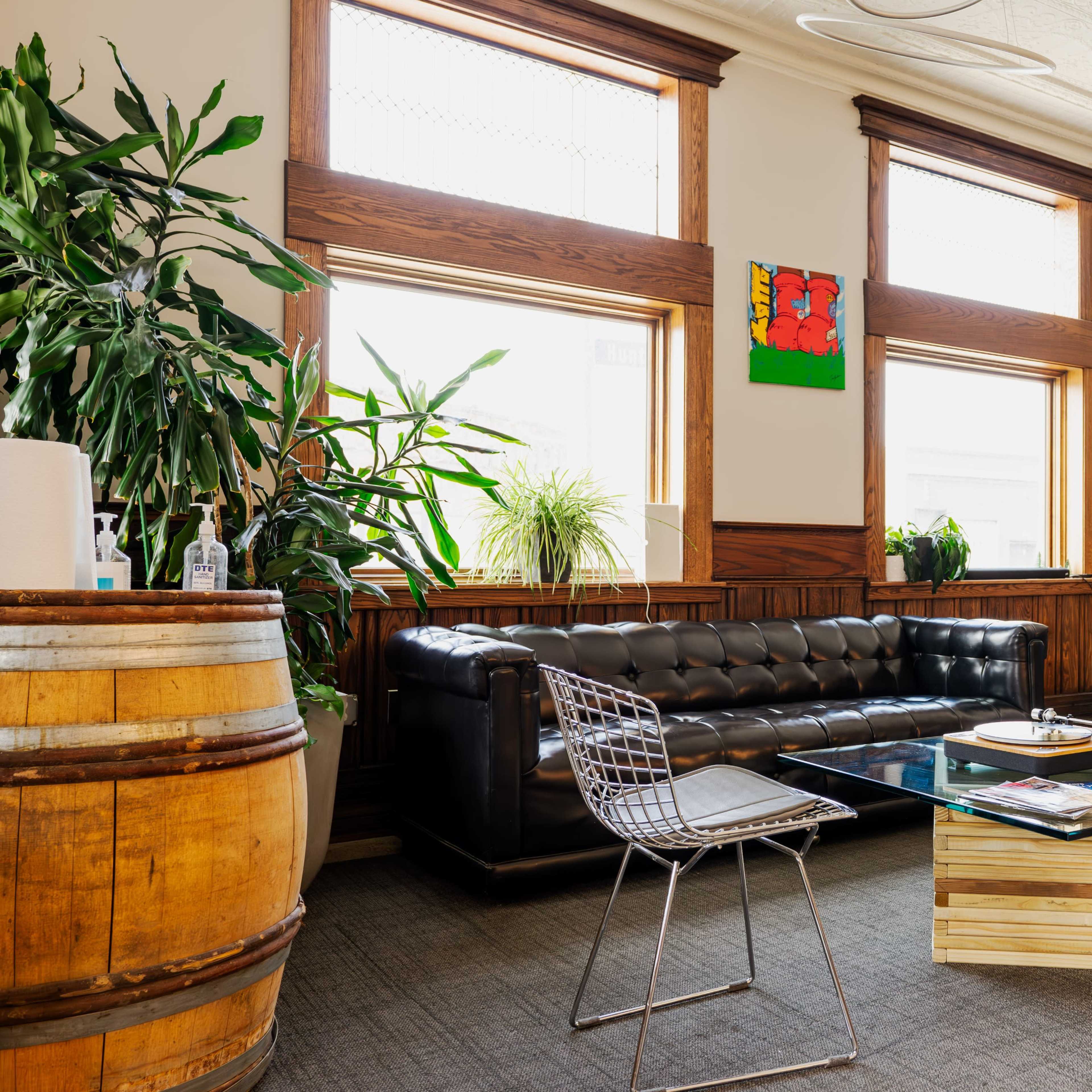 A modern lounge area featuring a black leather couch, a glass coffee table, wire chairs, and large windows adorned with plants and artwork.