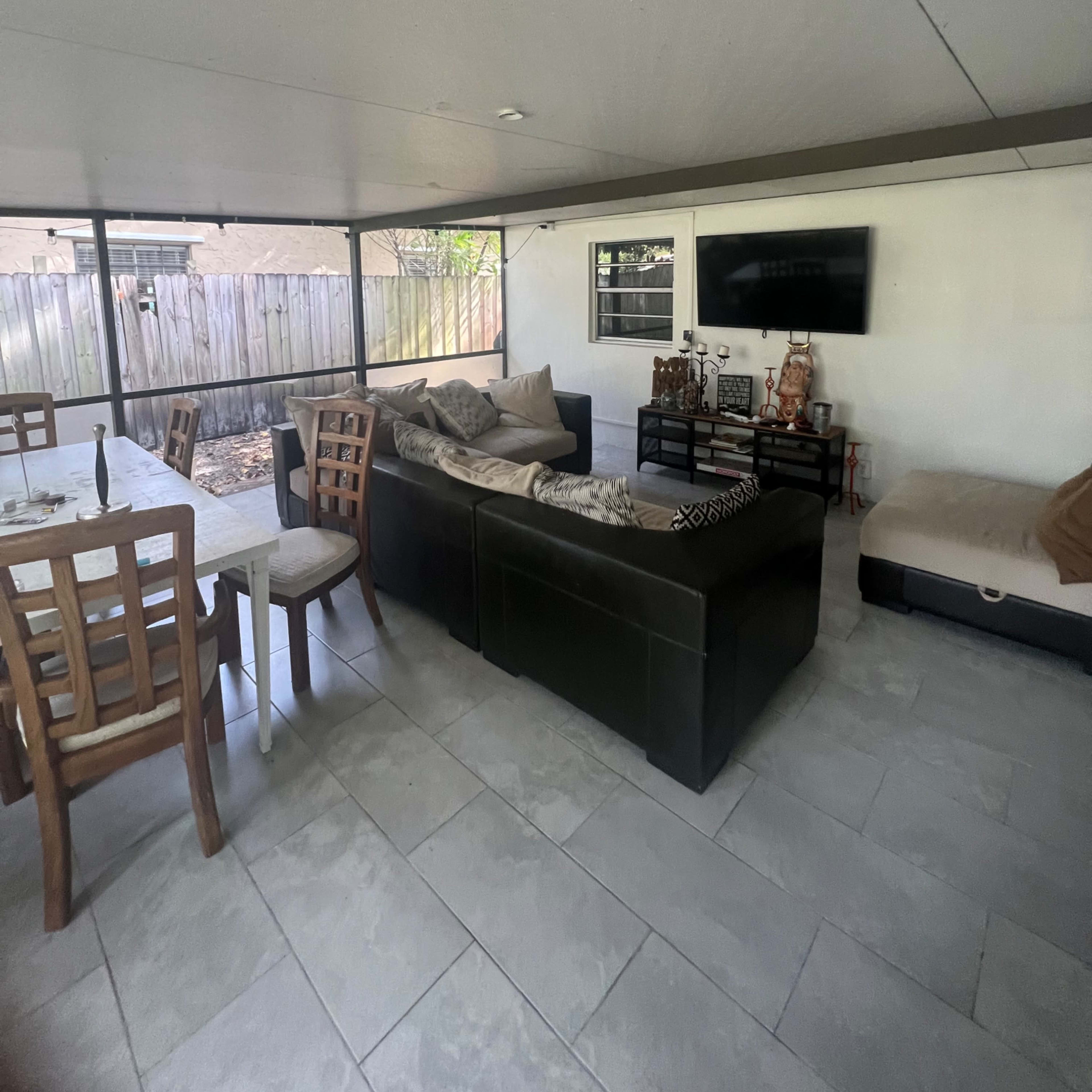 A covered outdoor living space featuring a dining table and chairs, a sofa with cushions, and a television mounted on the wall.