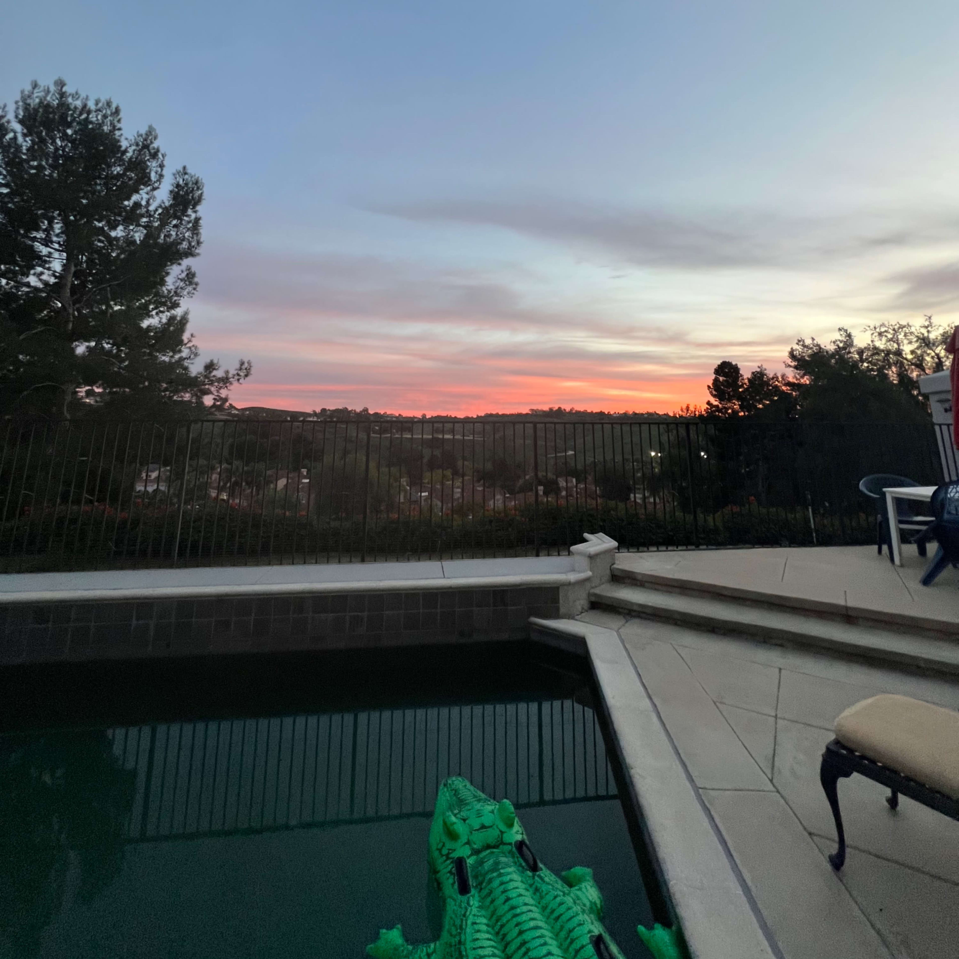 A poolside view at dusk with a green inflatable crocodile floating in the water and a sunset illuminating the sky.