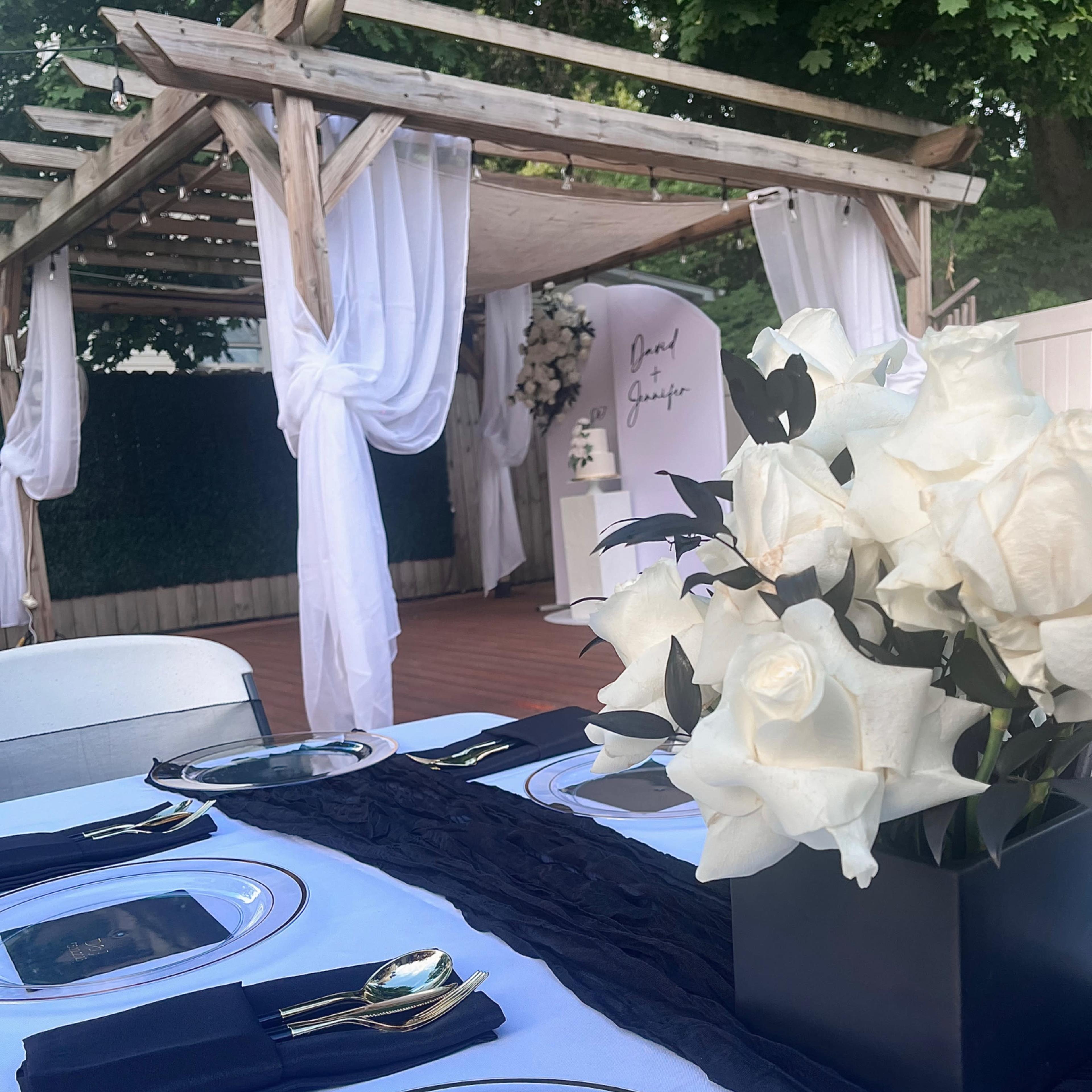 A beautifully arranged outdoor dining setup featuring a black table runner, white plates, and a centerpiece of white roses, with a draped pergola in the background.