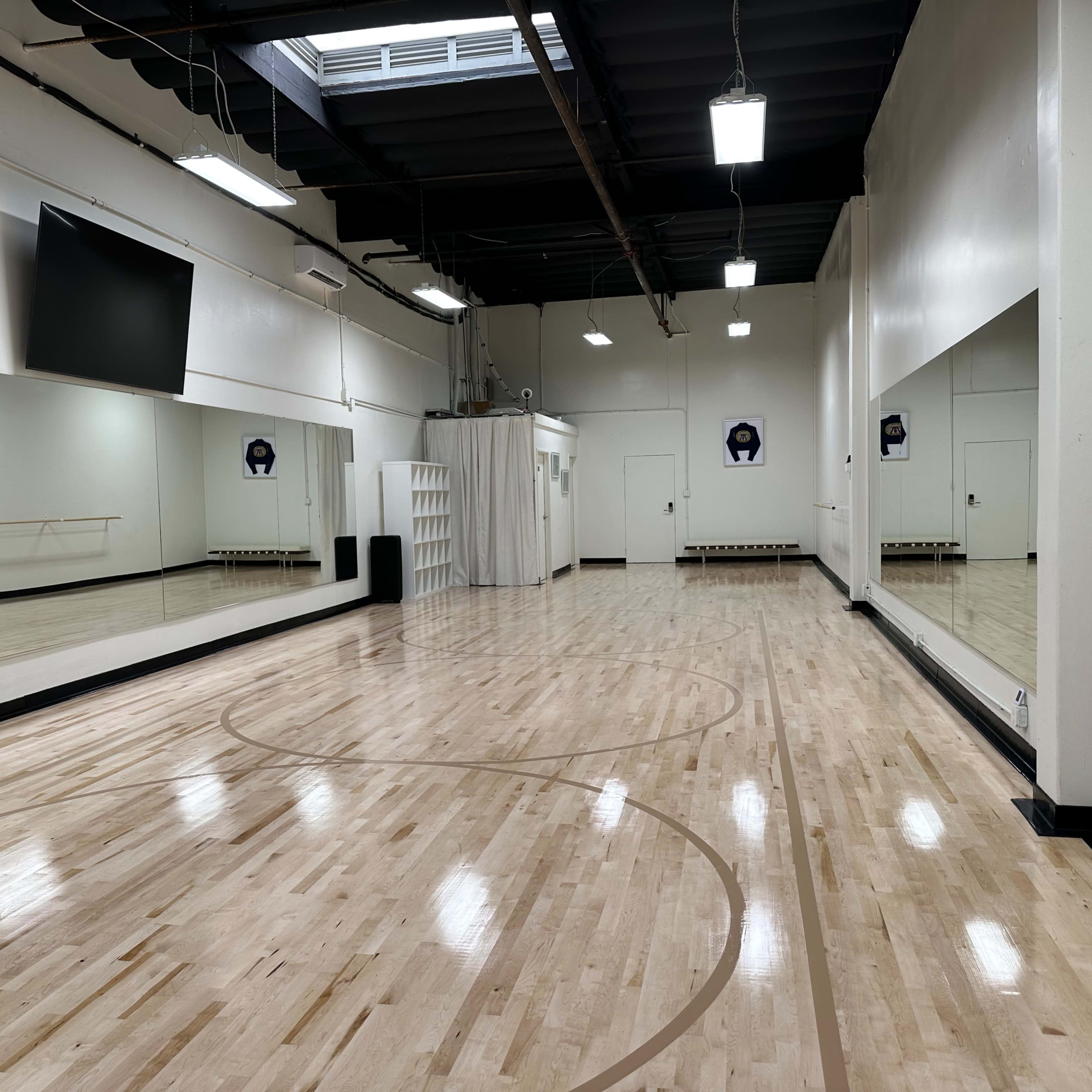 A spacious dance studio with wooden floors, large mirrors on the walls, and a mounted television.