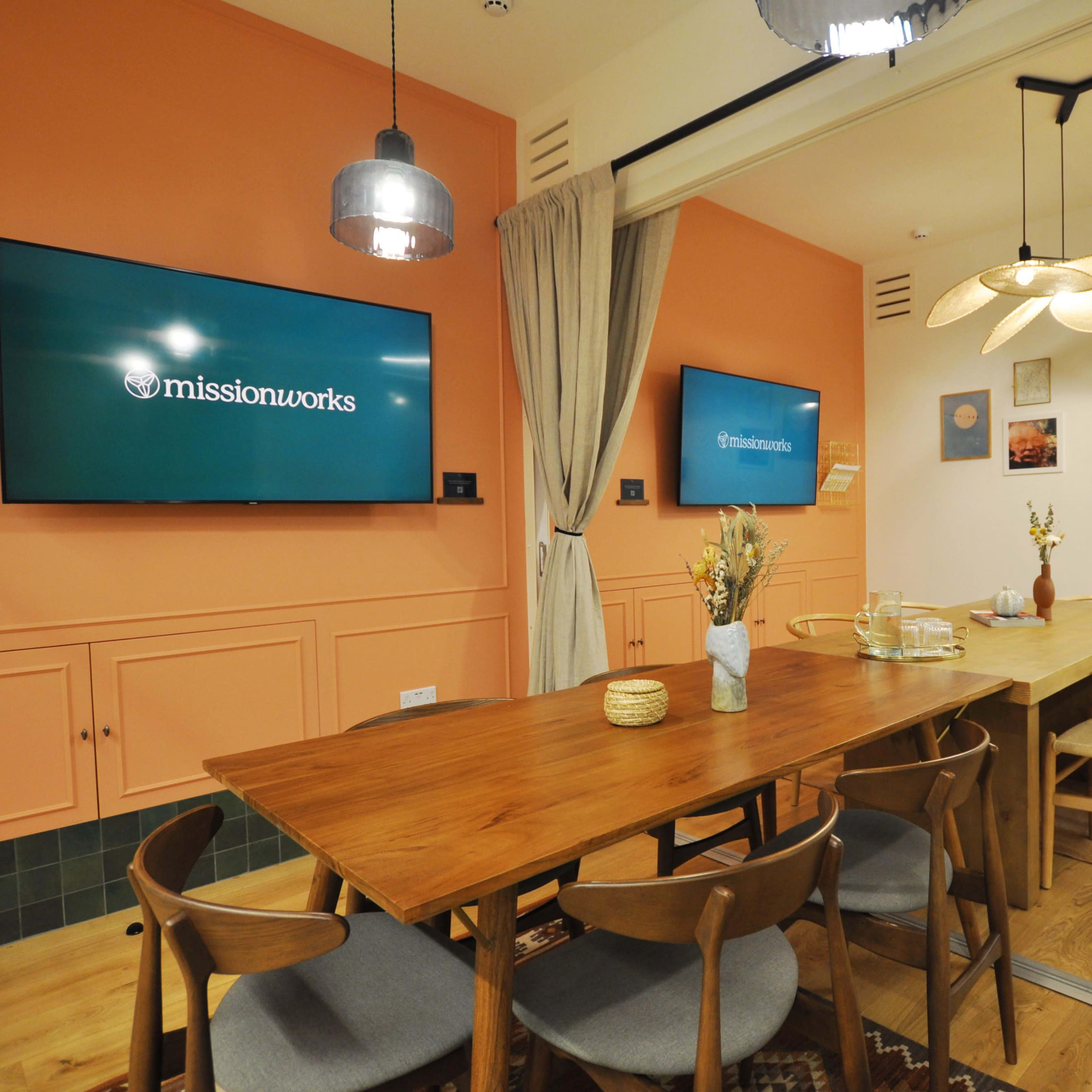A modern conference room with a long wooden table surrounded by chairs, featuring two wall-mounted screens and orange-painted walls.