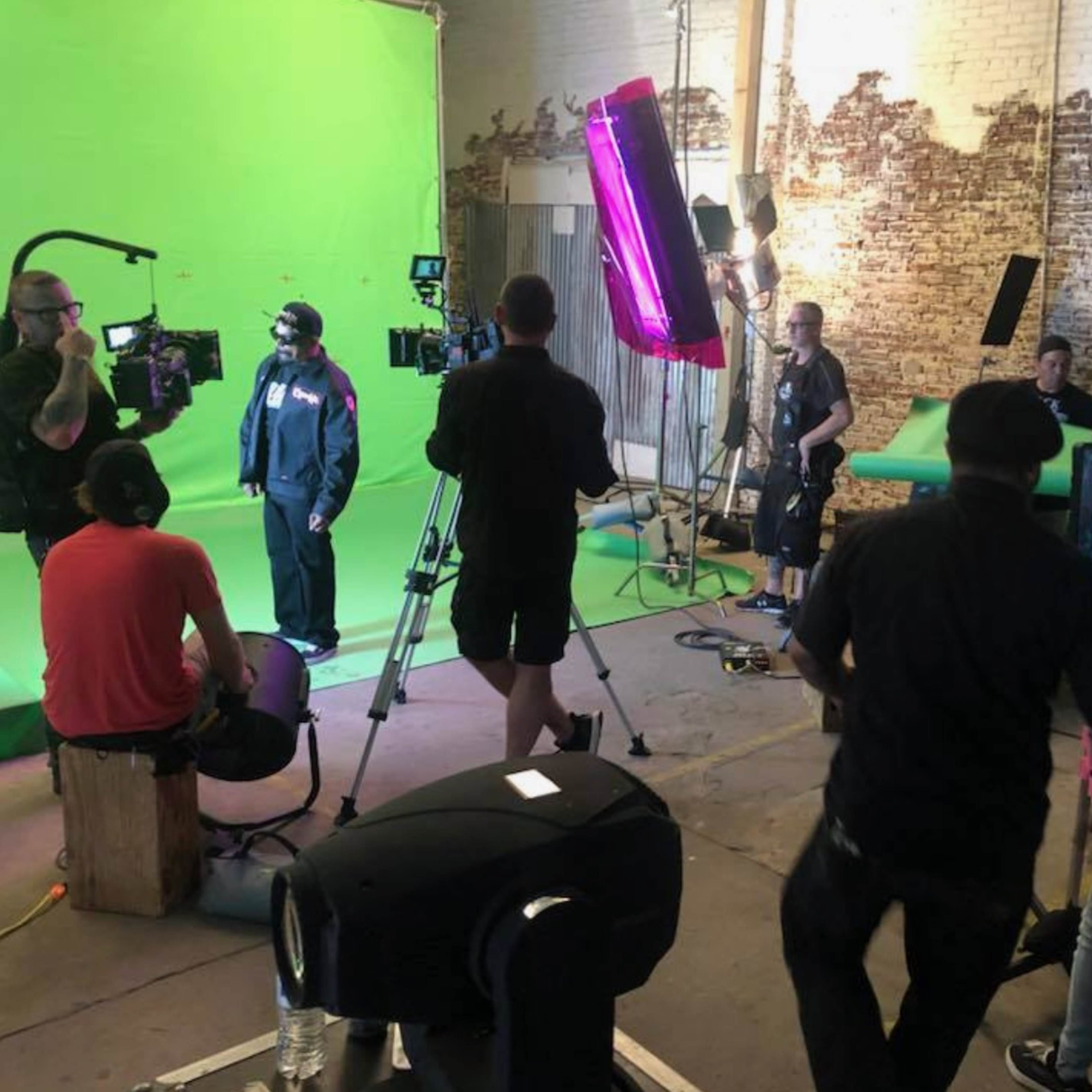 A film crew is set up in a studio with a green screen, multiple cameras, and lighting equipment.