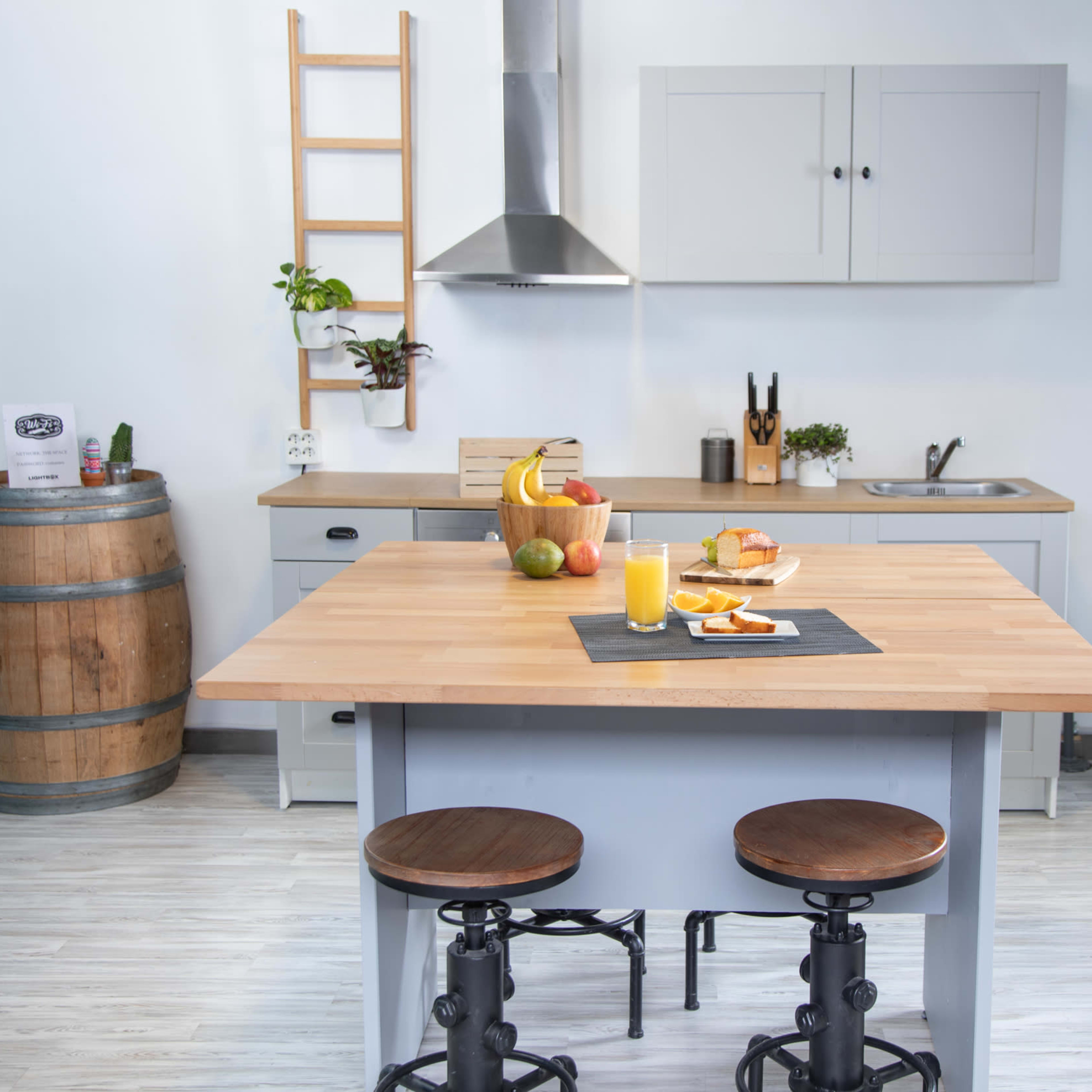 A modern kitchen features a wooden island with a variety of fruits and a drink, accompanied by two stools, and cabinets lined against a wall.