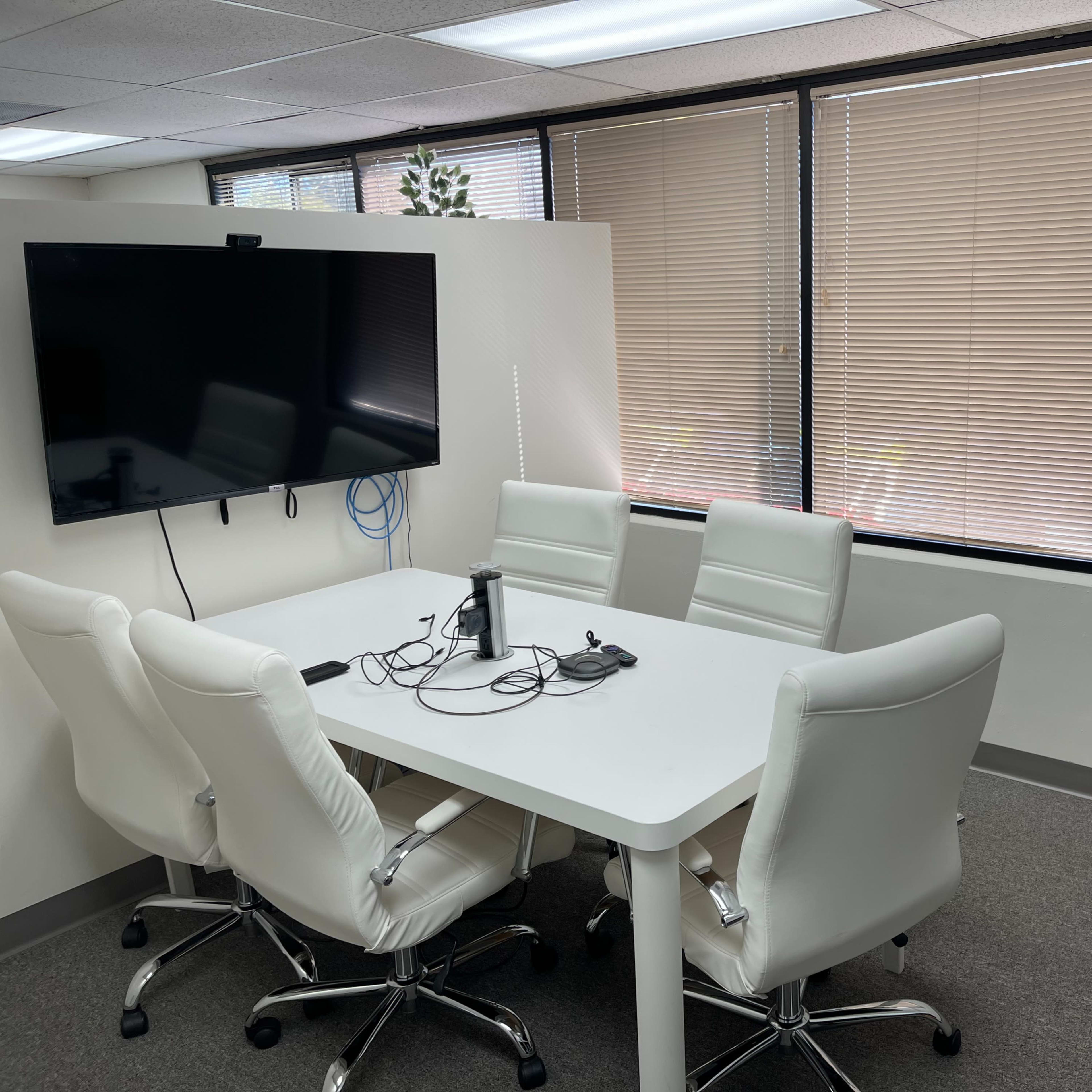A conference room features a white table surrounded by four white chairs, with a large screen mounted on the wall and natural light coming through window blinds.