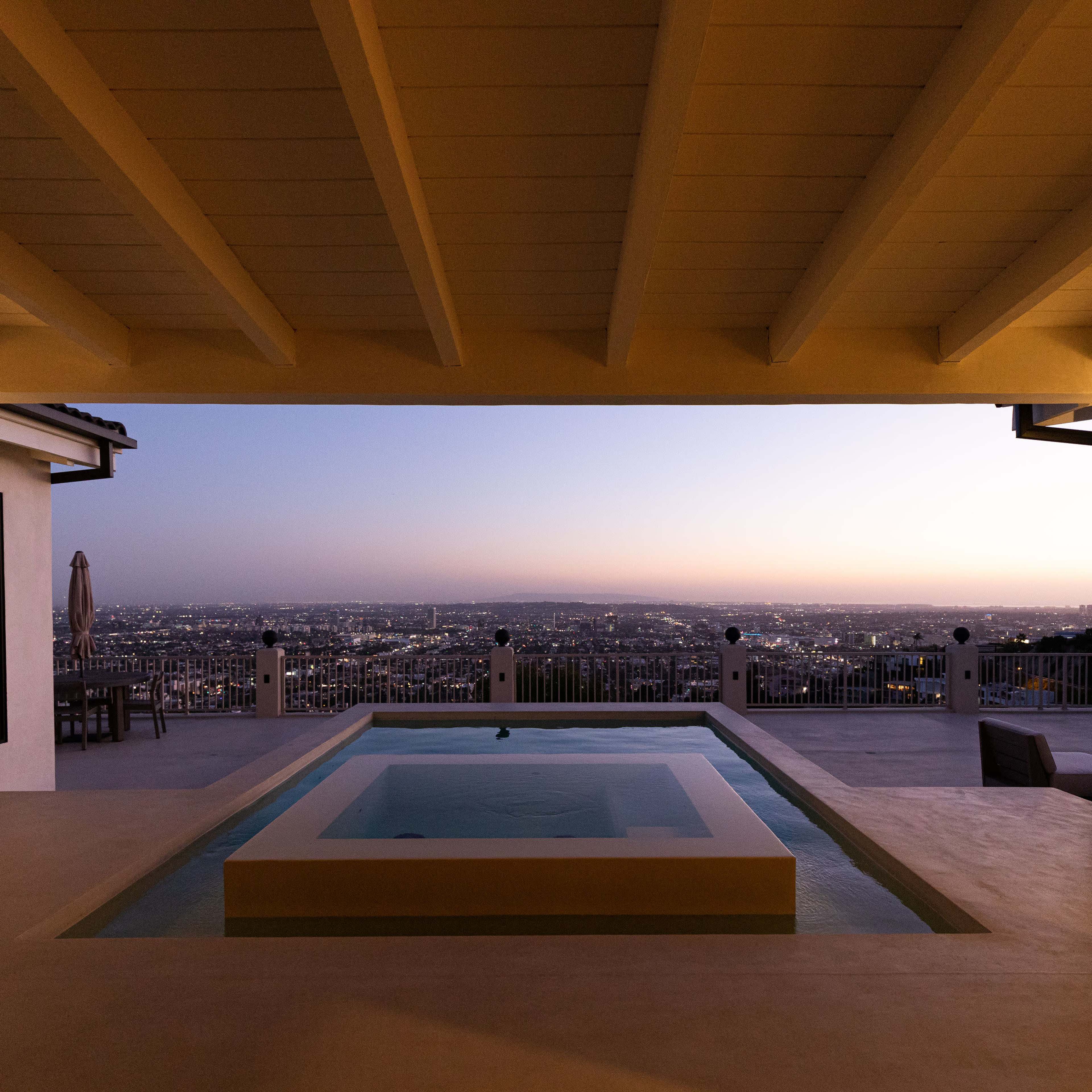 A modern outdoor space with a pool overlooking a cityscape at dusk, framed by a spacious overhang.