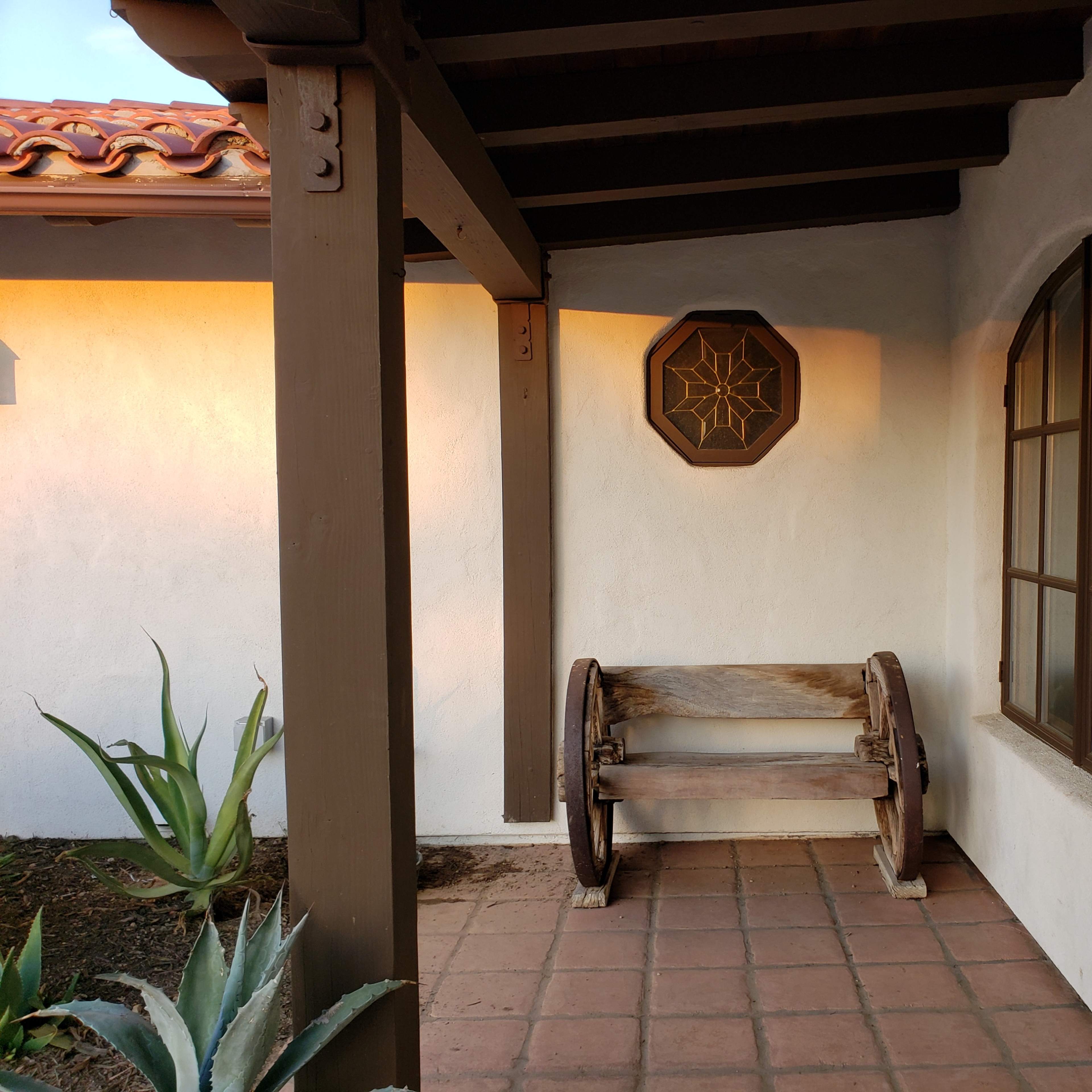 A quaint porch with a wooden bench, a decorative wall piece, and a lantern, surrounded by plants and tiled flooring.