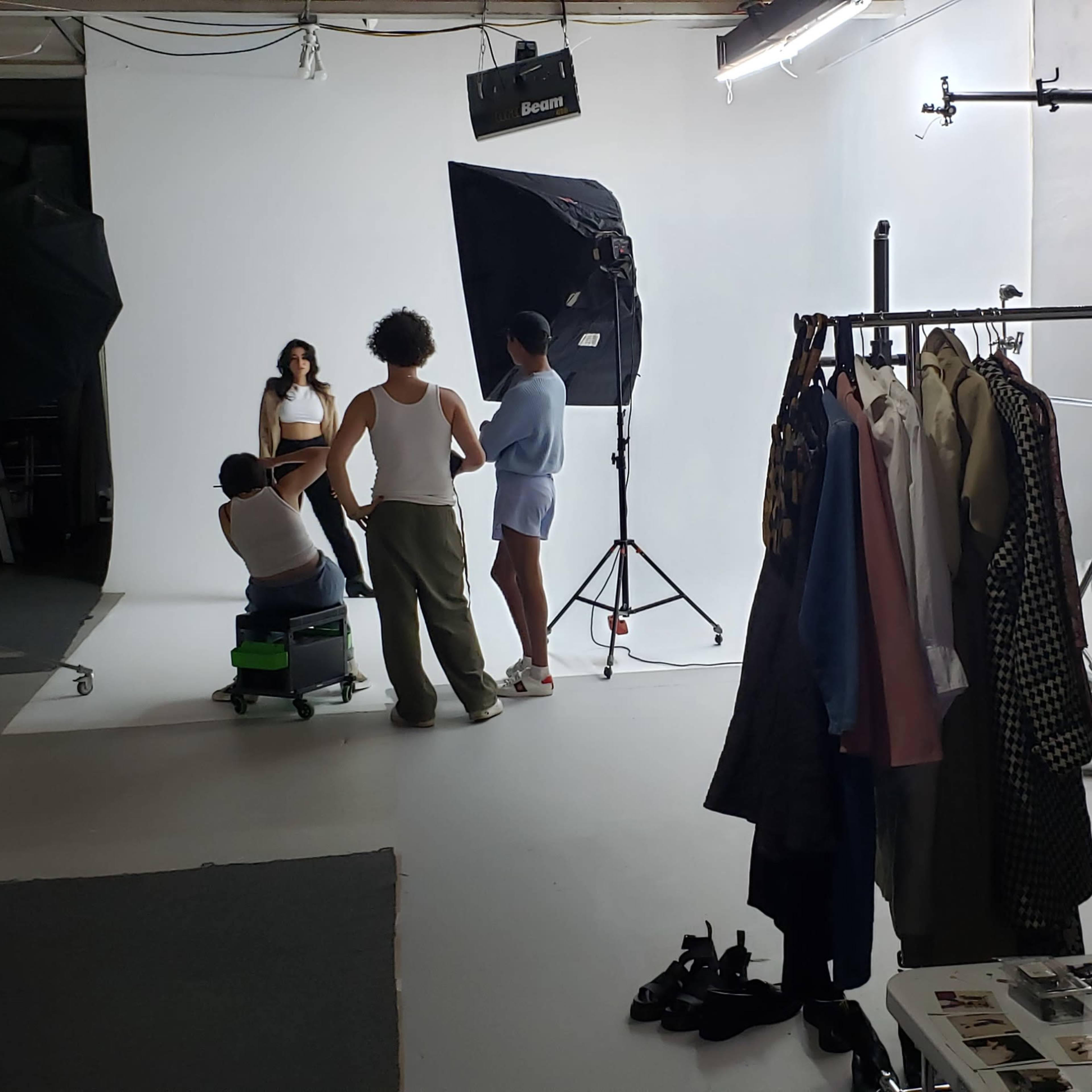 A group of people are engaged in a photoshoot in a well-equipped studio, with clothing racks and various photography equipment visible.