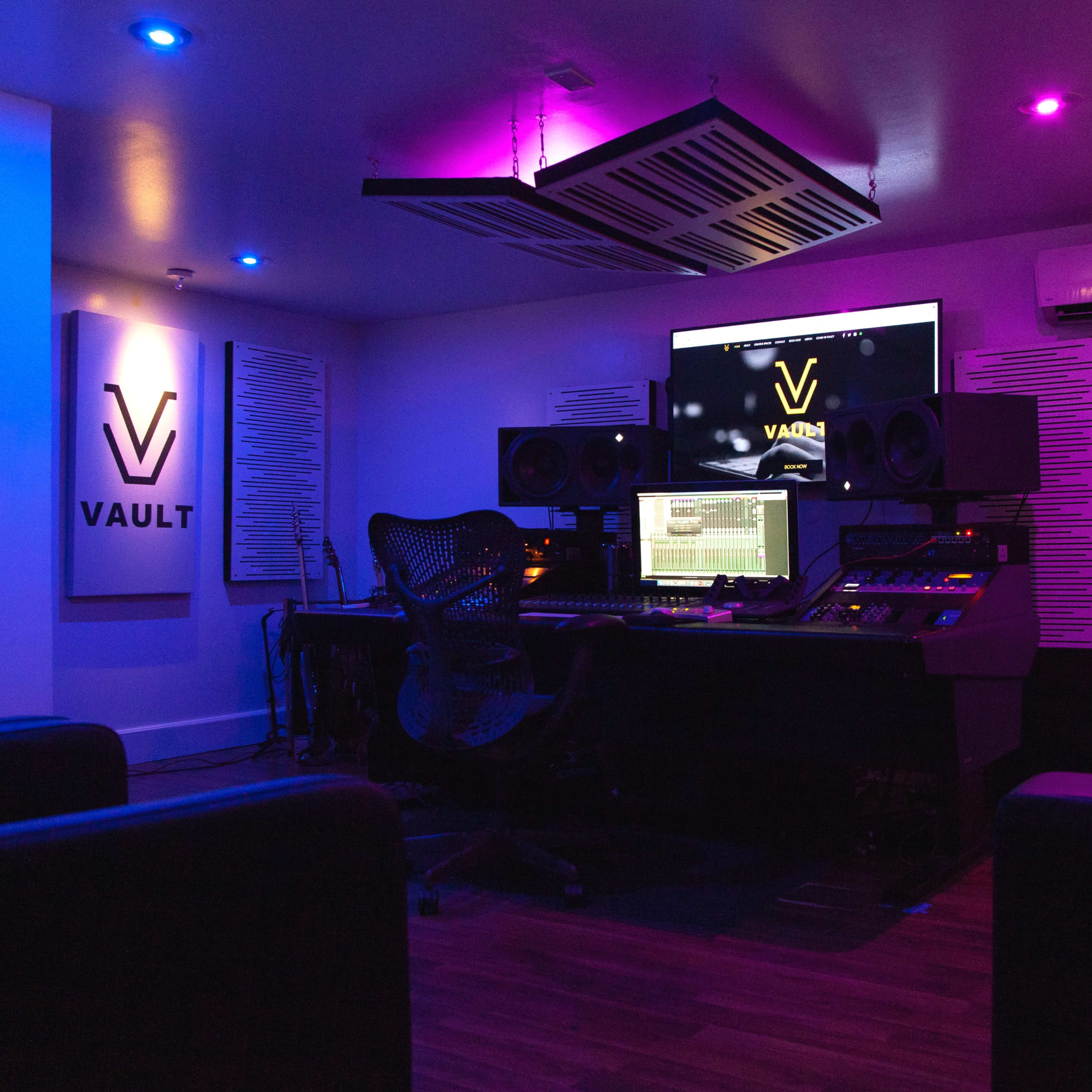 A modern recording studio with a dark ambiance, featuring a large mixing console, multiple speakers, and colorful LED lighting.