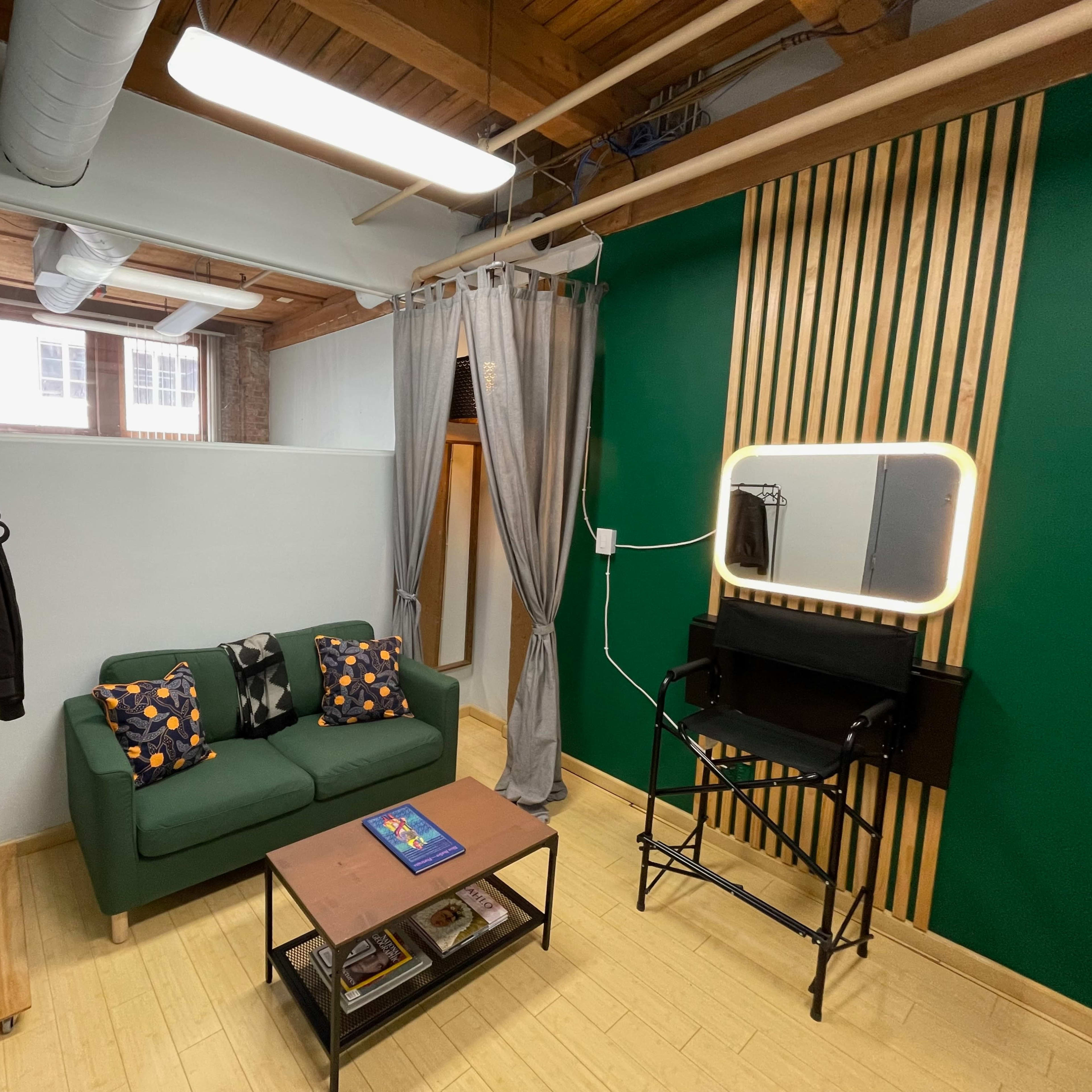 A small room with a green accent wall, a green sofa, a black director's chair, a mirror with lights, and a wooden coffee table.