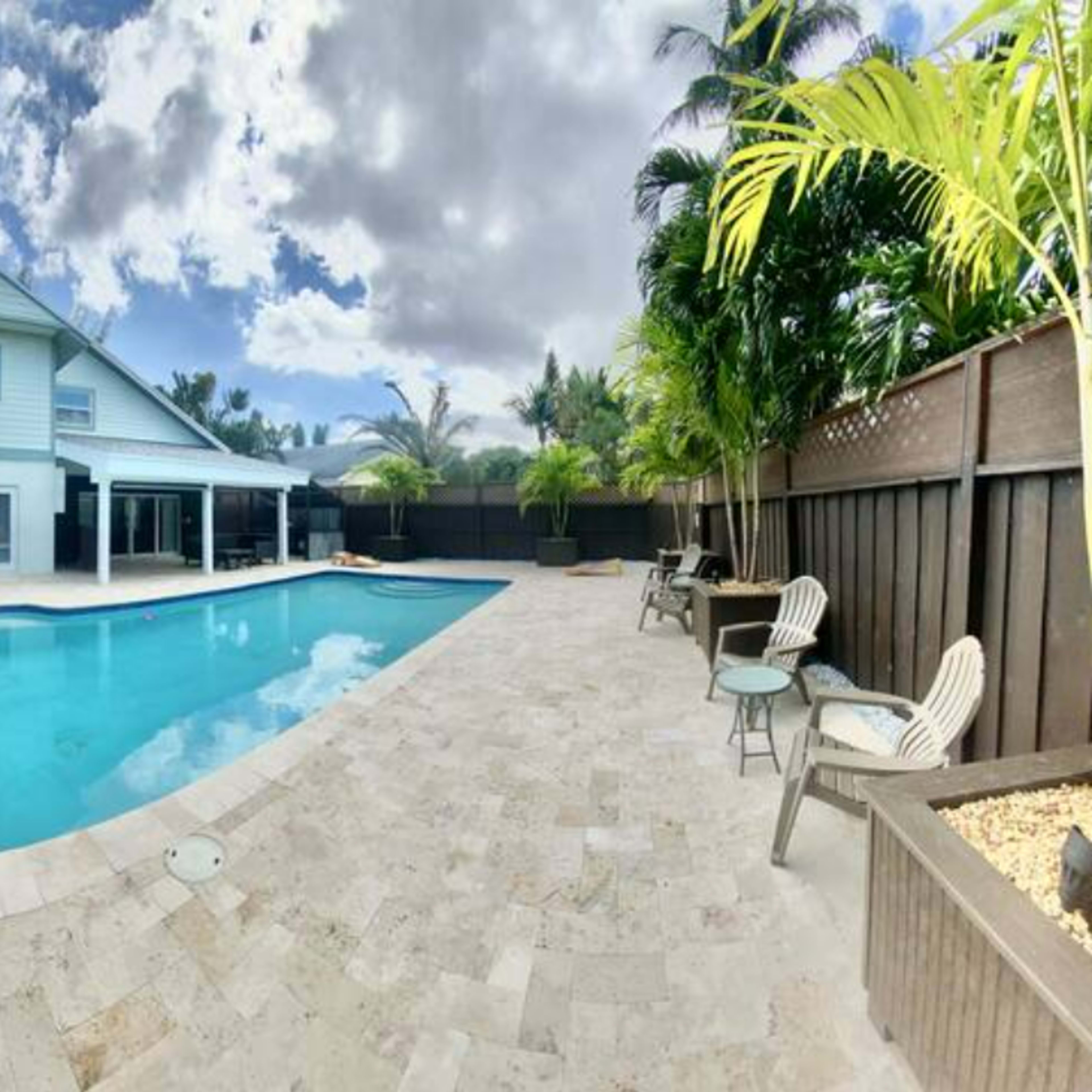 A rectangular swimming pool surrounded by lounge chairs and tropical plants, with a two-story house in the background under a partly cloudy sky.