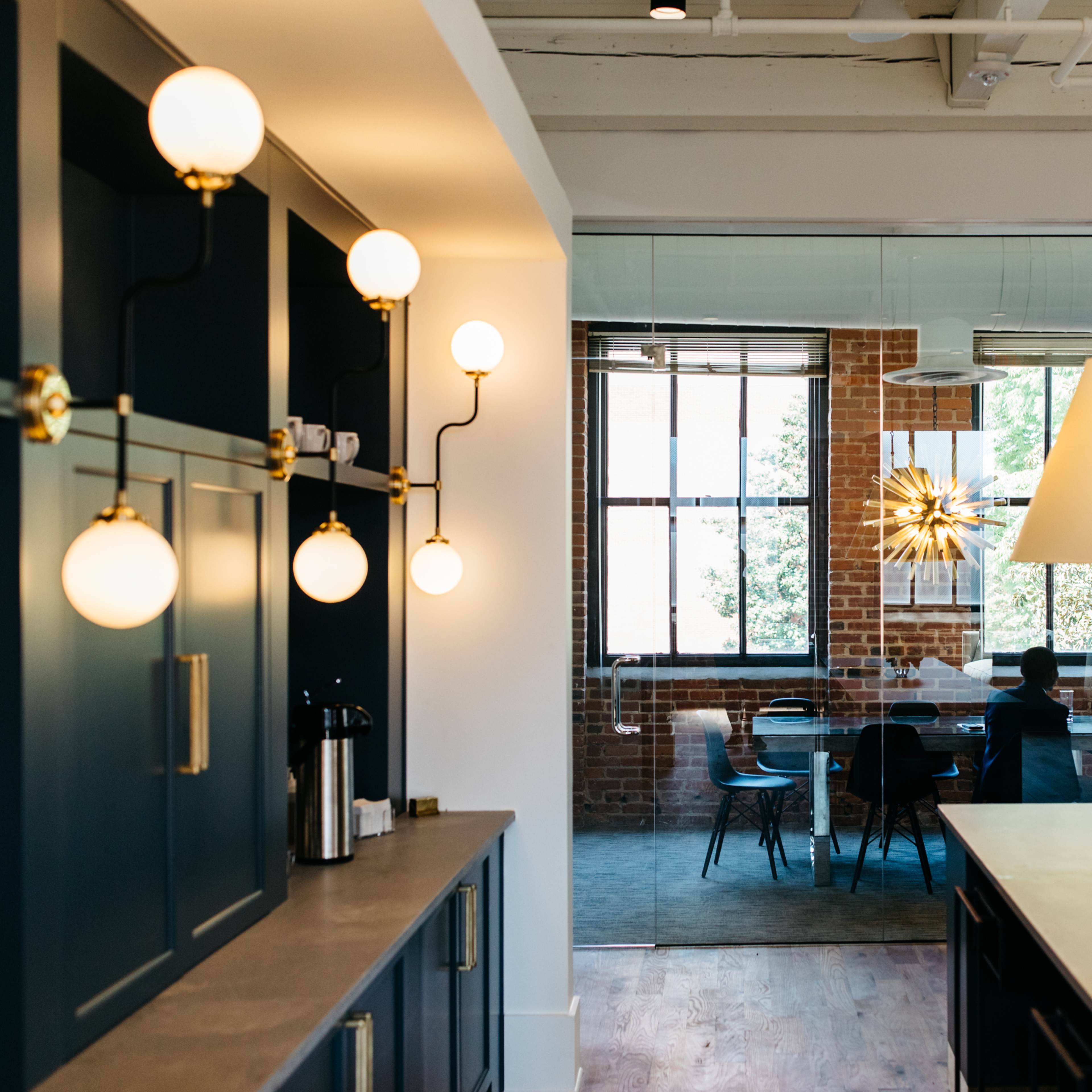A modern office space with a kitchen area featuring round light fixtures and a dining table visible through a glass partition.