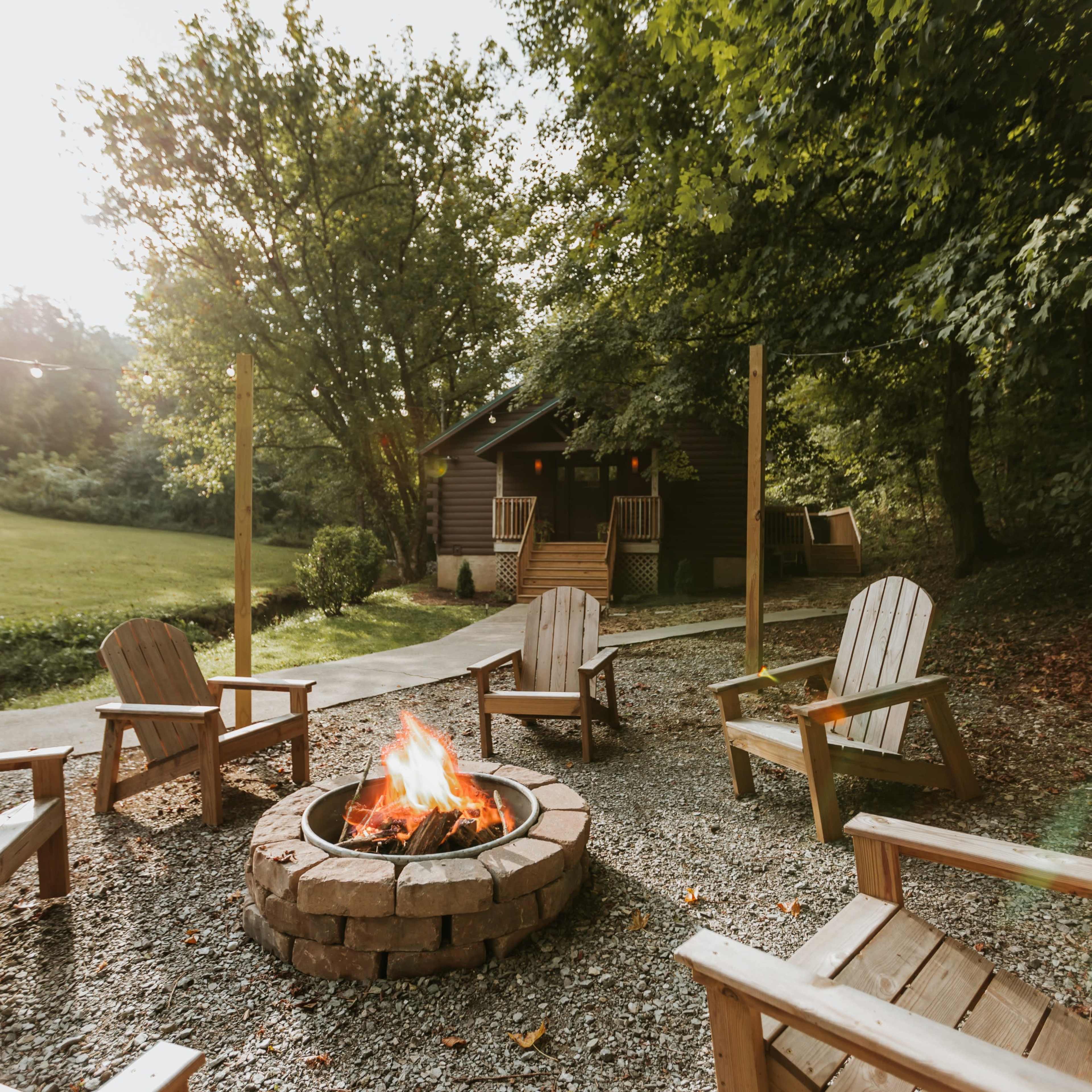 A fire pit surrounded by wooden chairs is set in a clearing in front of a cabin, with trees and a grassy area in the background.