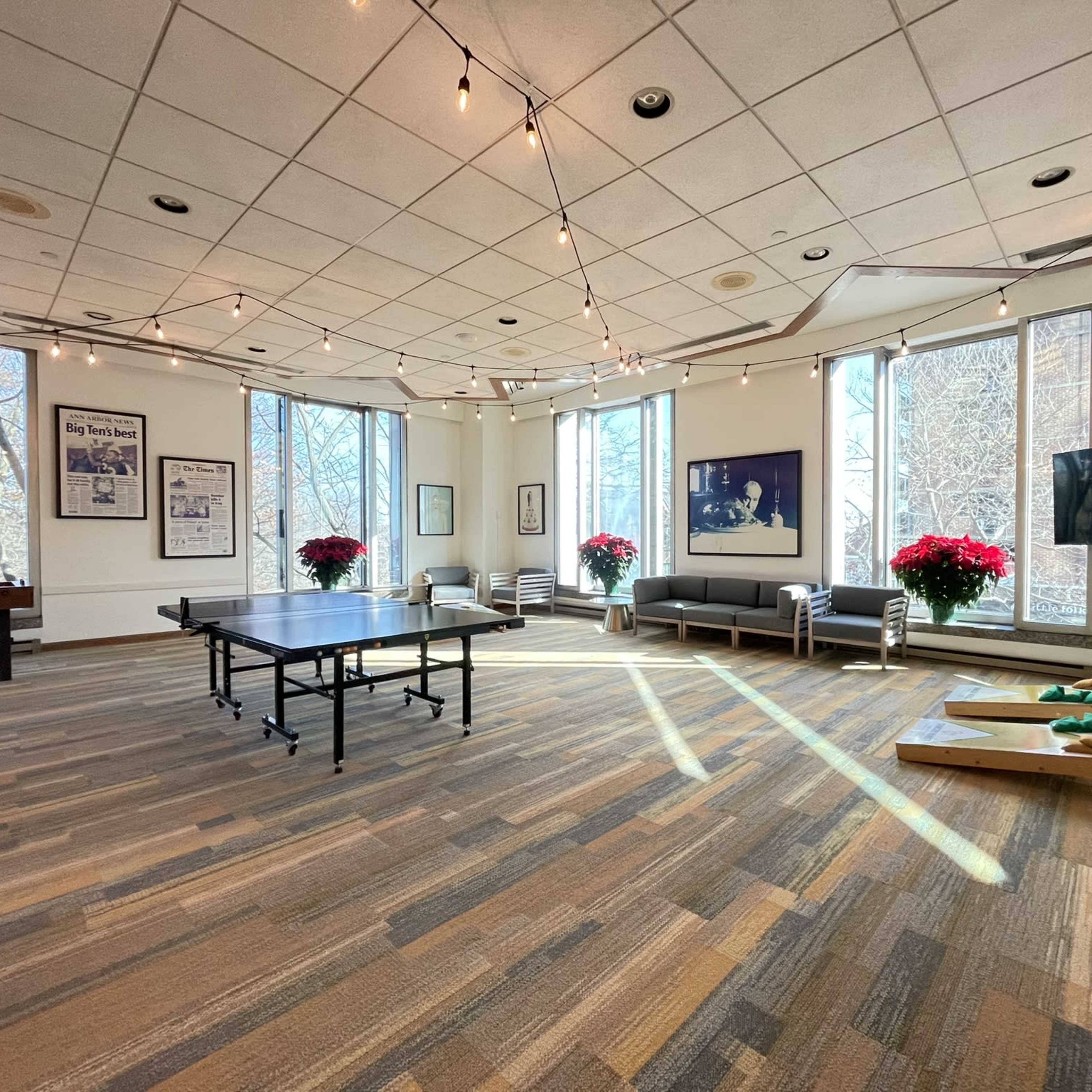 A spacious room with large windows, string lights overhead, a ping pong table, and a seating area adorned with potted plants.