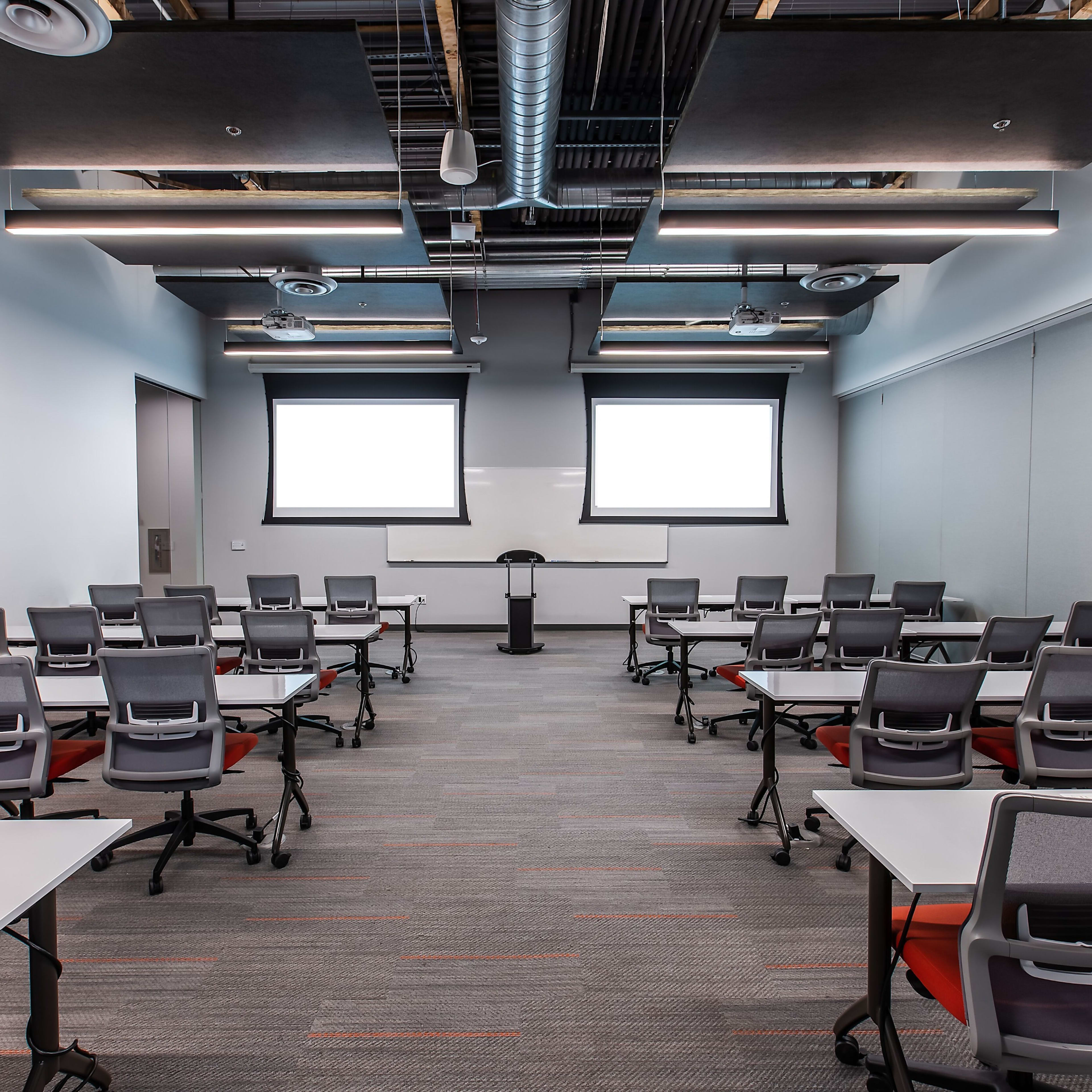 A training room setup with rows of tables and chairs facing a podium and two large projection screens.