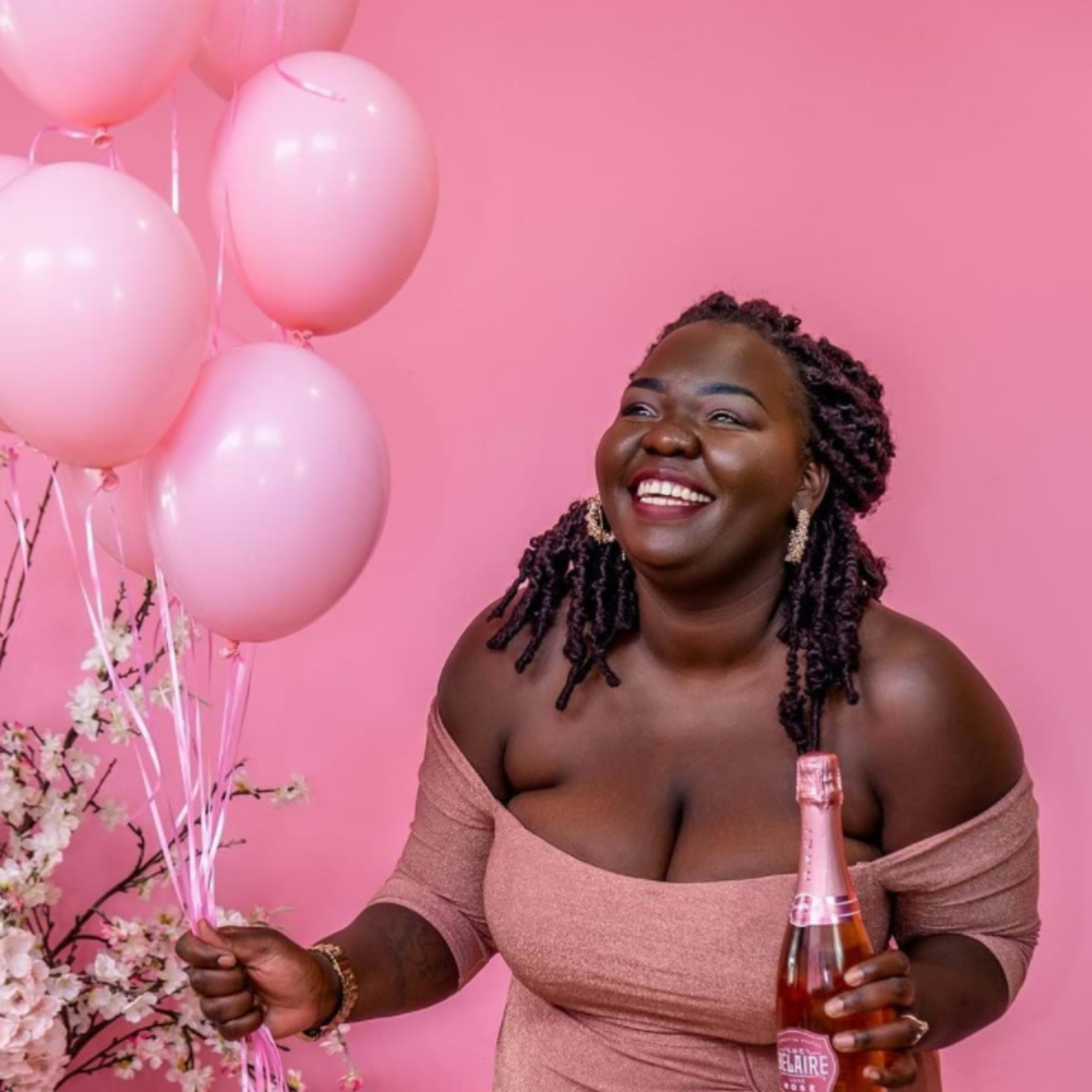 A woman smiles while holding a bottle of sparkling wine and pink balloons against a pink backdrop.