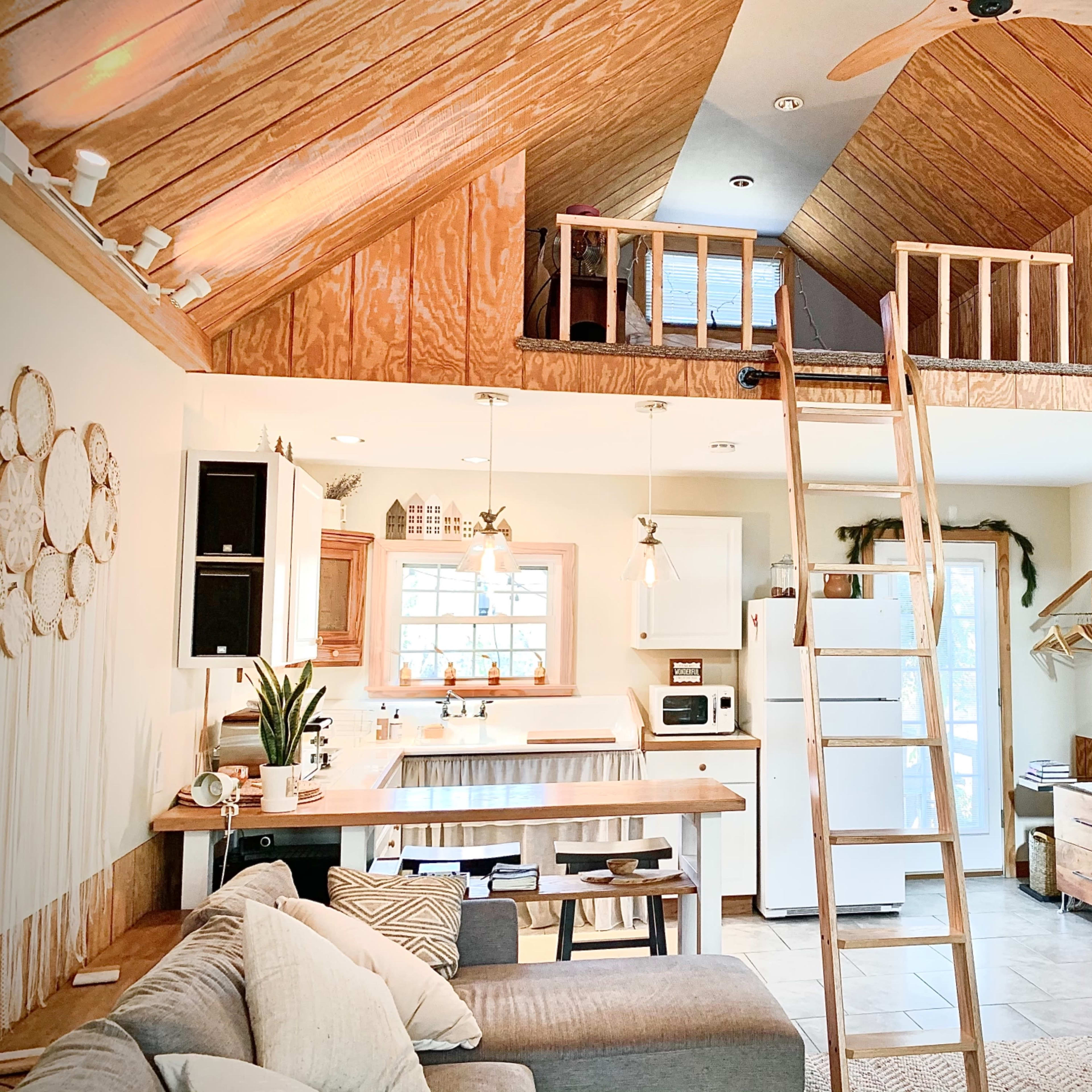A cozy living space featuring a loft area, wooden beams, a kitchen with a small island, and a staircase leading to the loft.