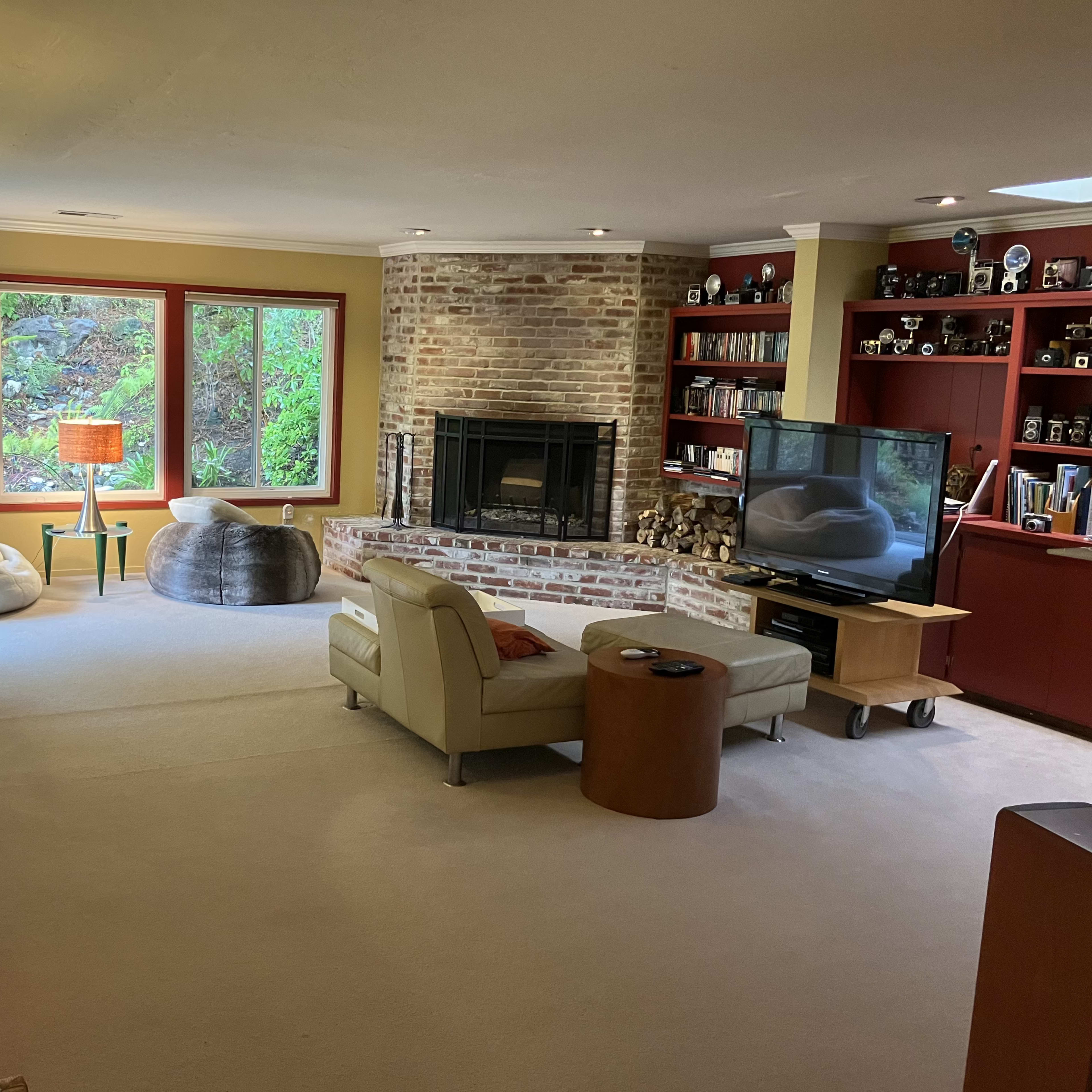 The living room features a pair of bean bag chairs, a brick fireplace, a television on a wheeled cart, and large windows with views of greenery outside.