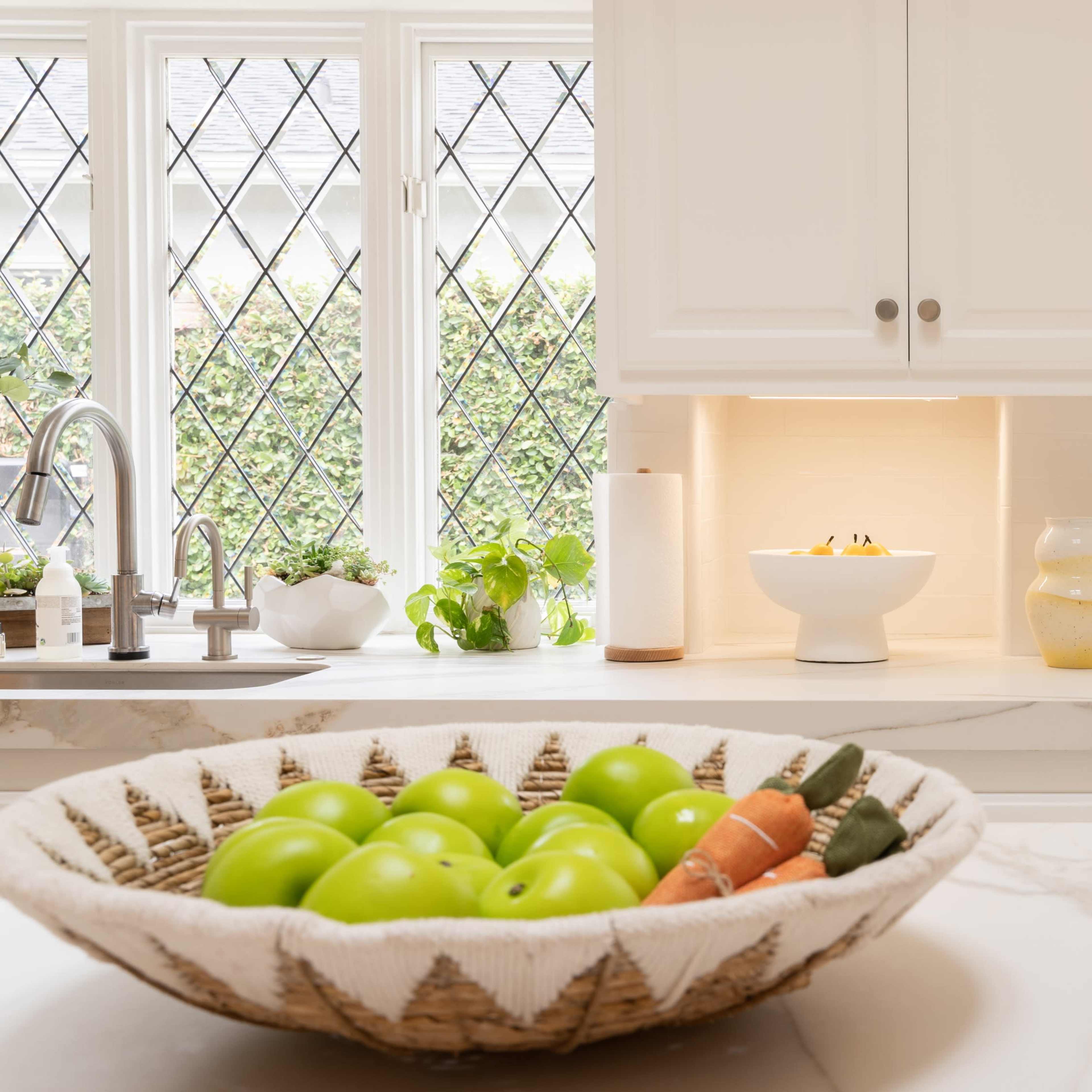 A kitchen scene features a bowl of green apples and carrots on a marble countertop, with large windows and a door showing a geometric glass pattern.