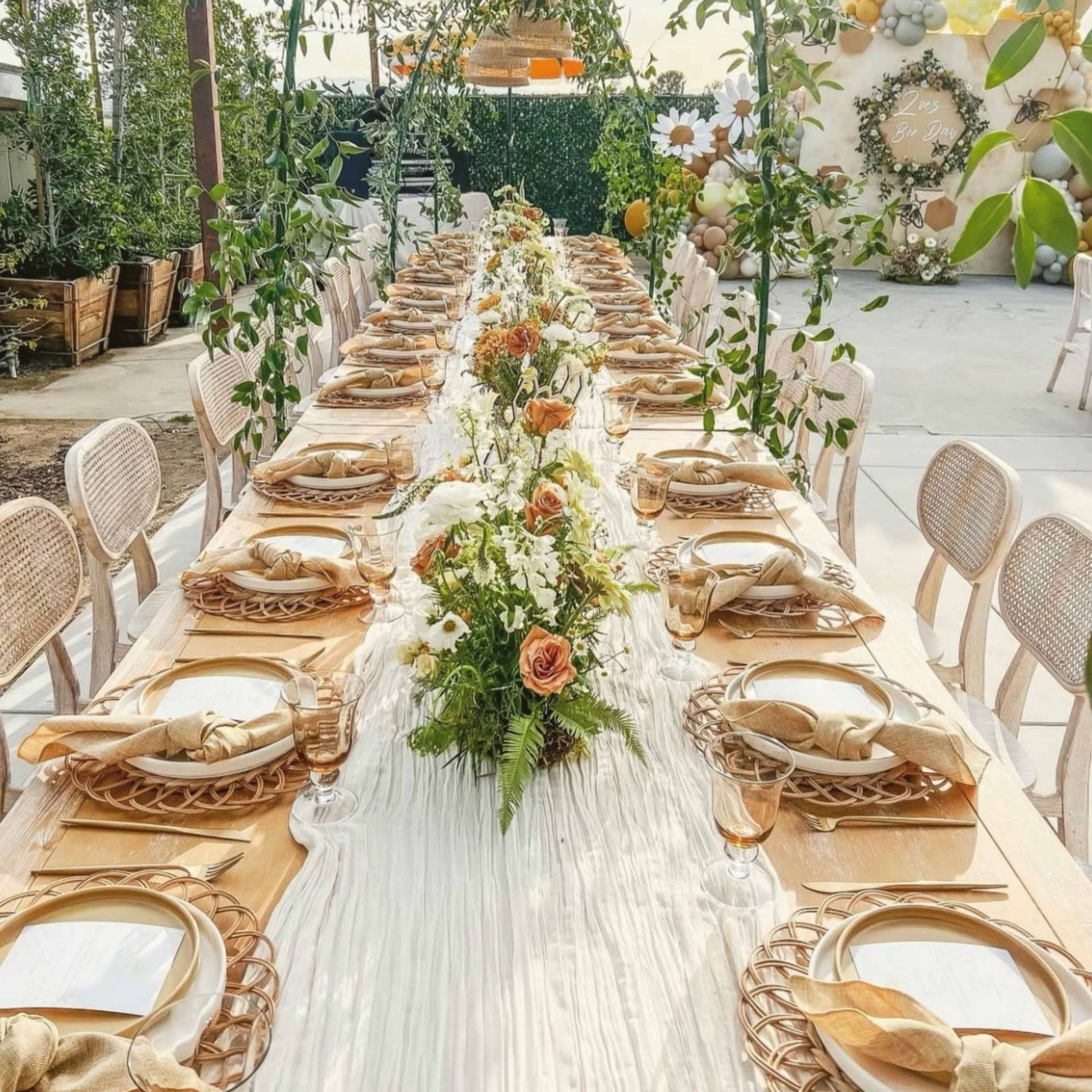 A long, elegantly set outdoor dining table is adorned with floral centerpieces and surrounded by greenery under hanging wicker light fixtures.