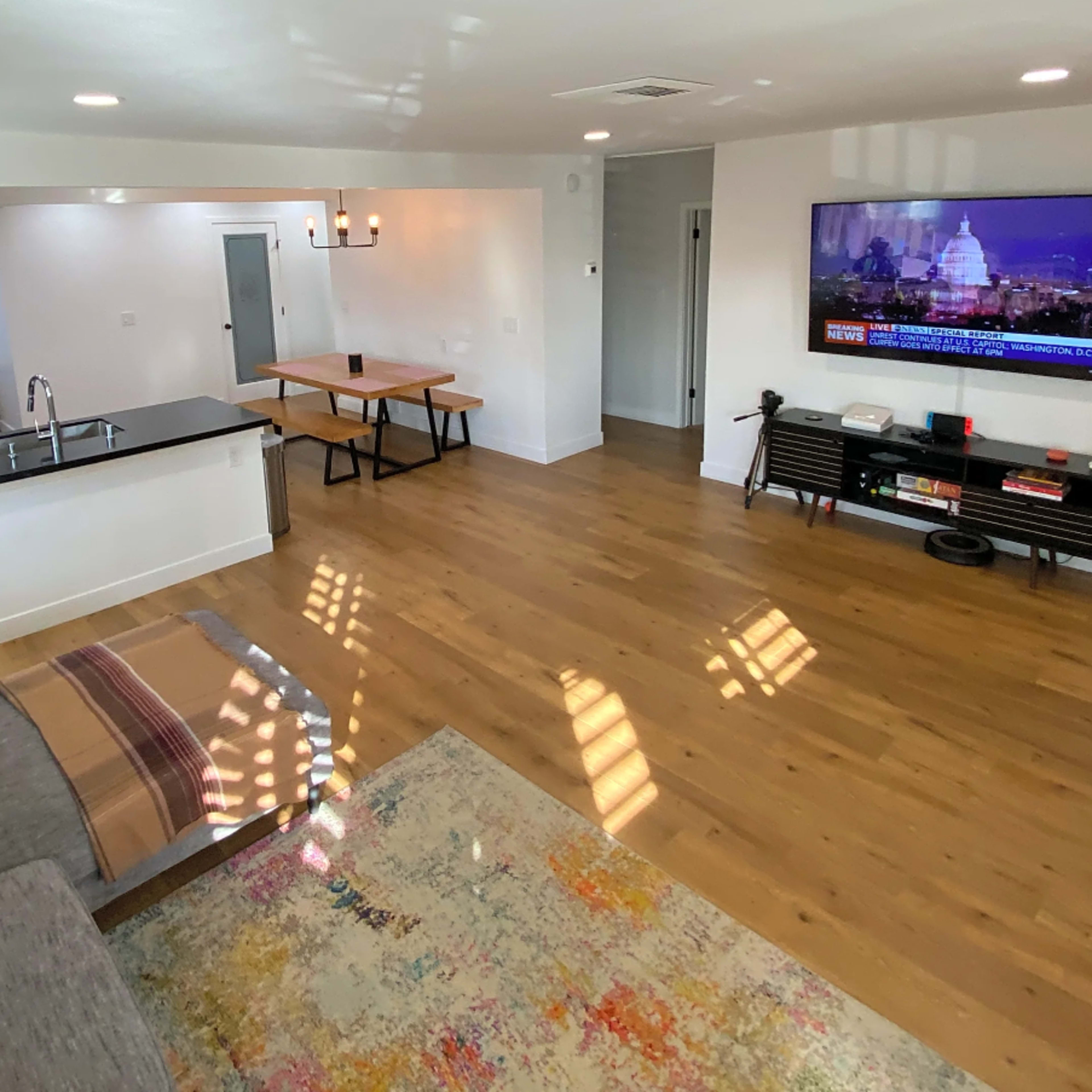 A modern living room features a gray sofa, a glossy black kitchen countertop, a wooden dining table, and a large TV displaying news.