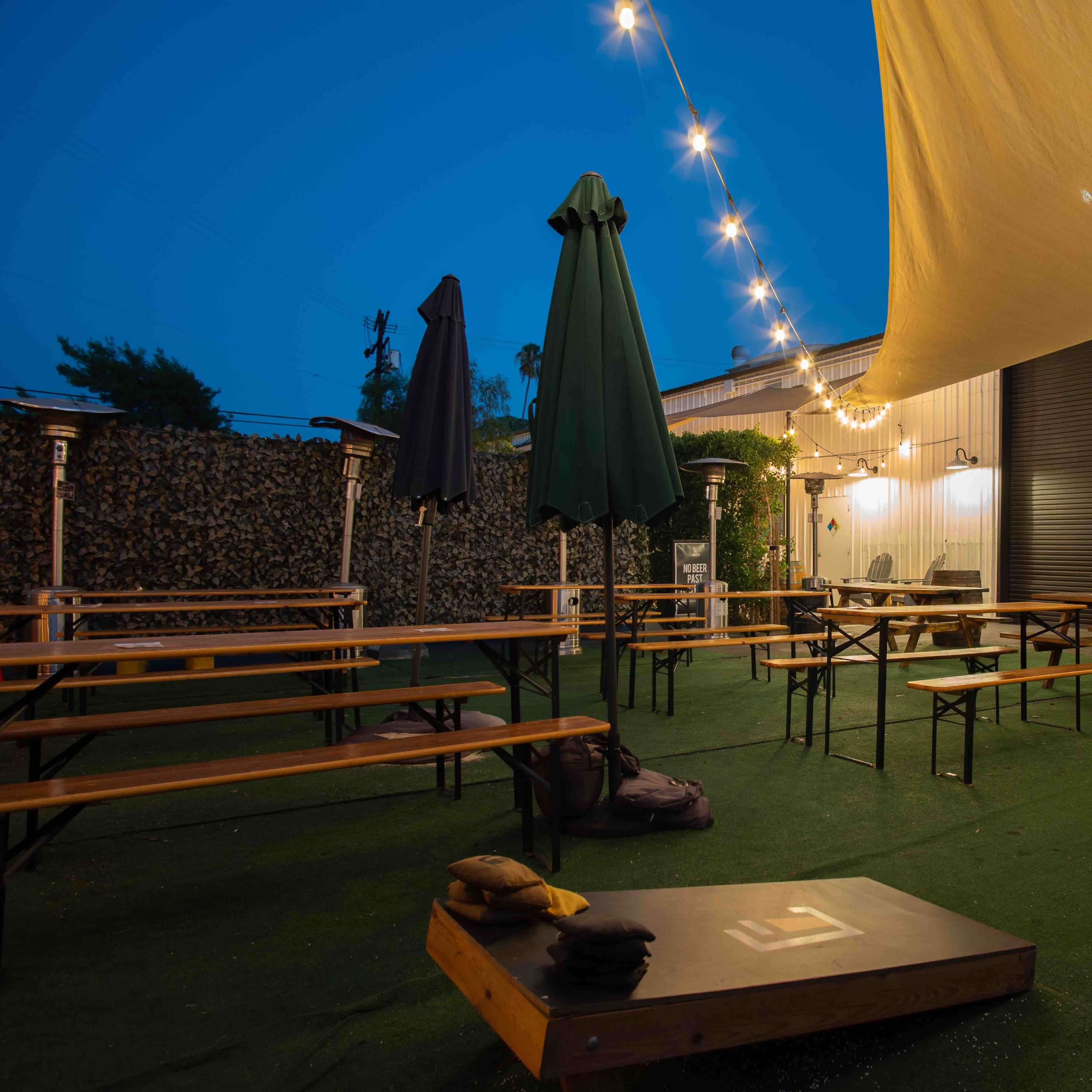 An outdoor seating area with wooden benches and tables, illuminated by string lights and decorated with greenery.