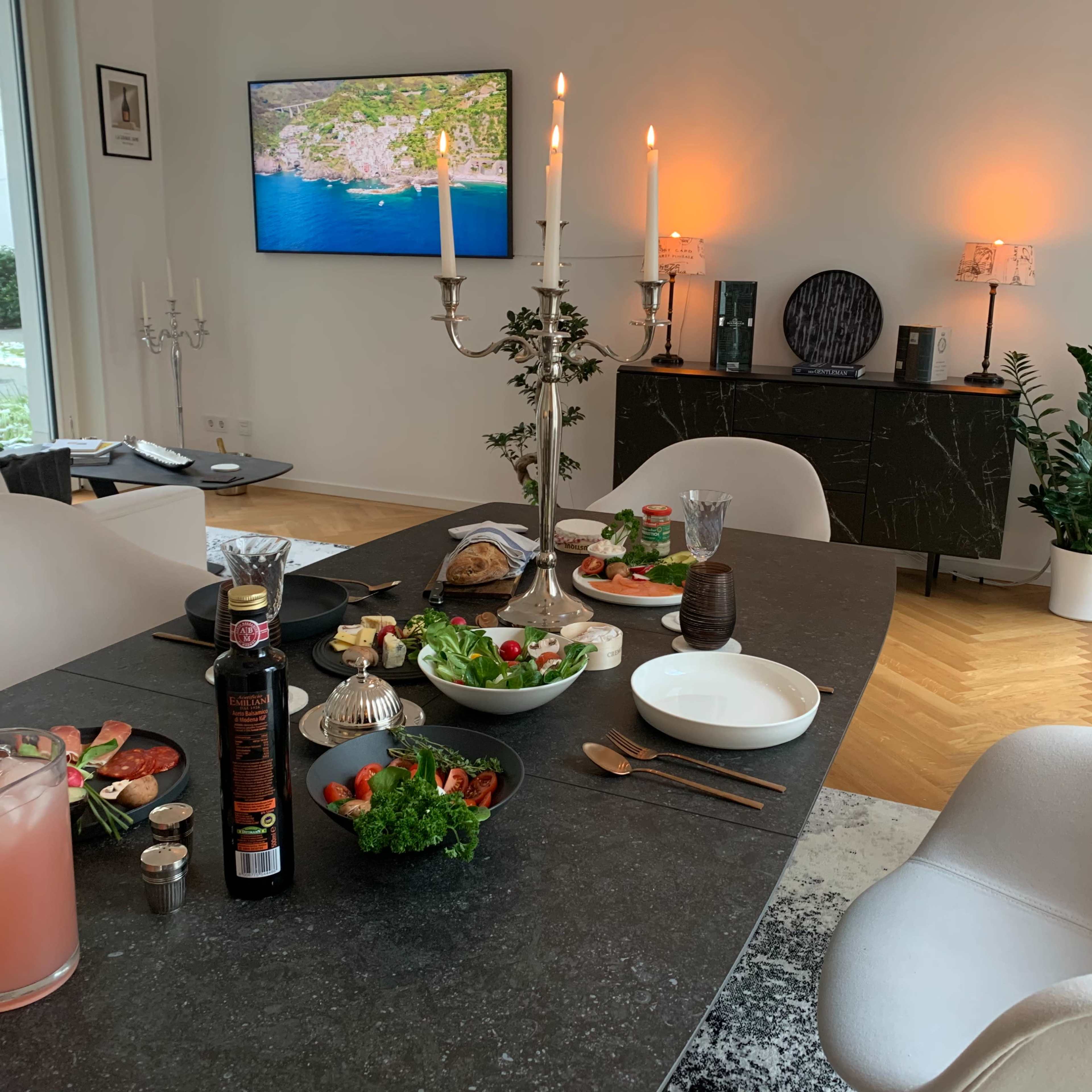 A dining table is set with food and drinks, while a modern living space includes a TV, decorative lighting, and potted plants.