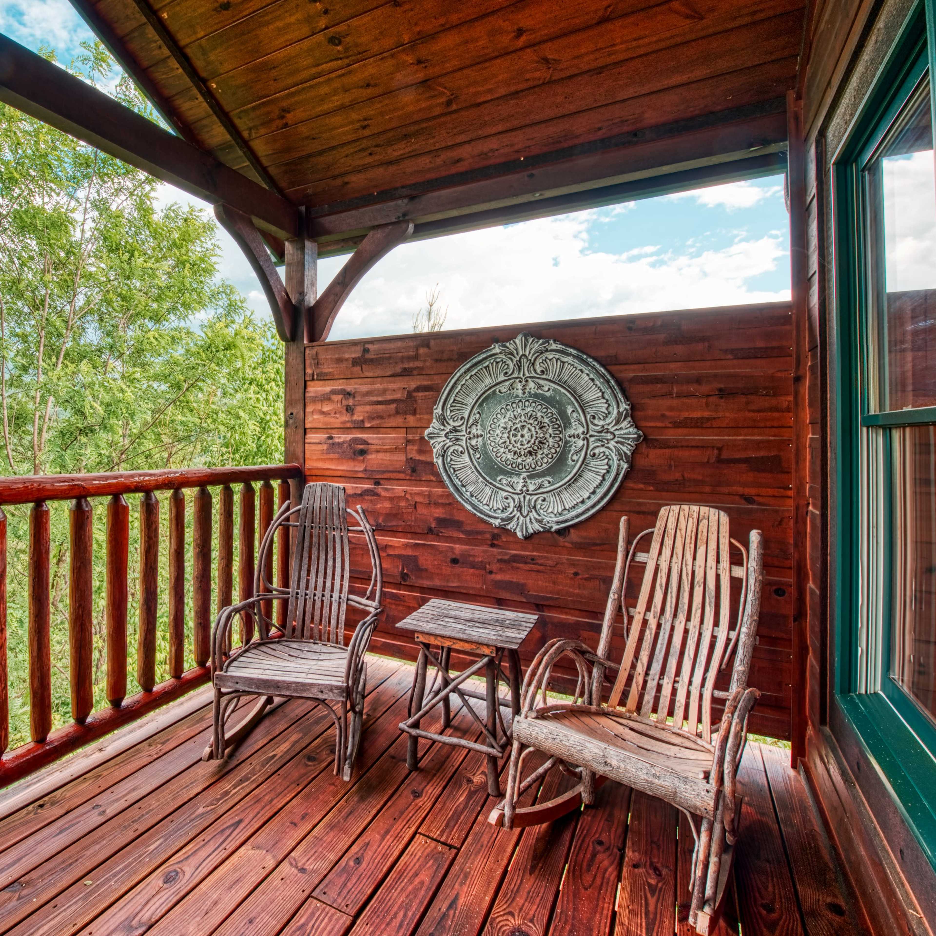 A wooden balcony with two rocking chairs and a small table, surrounded by trees and mountains in the background.