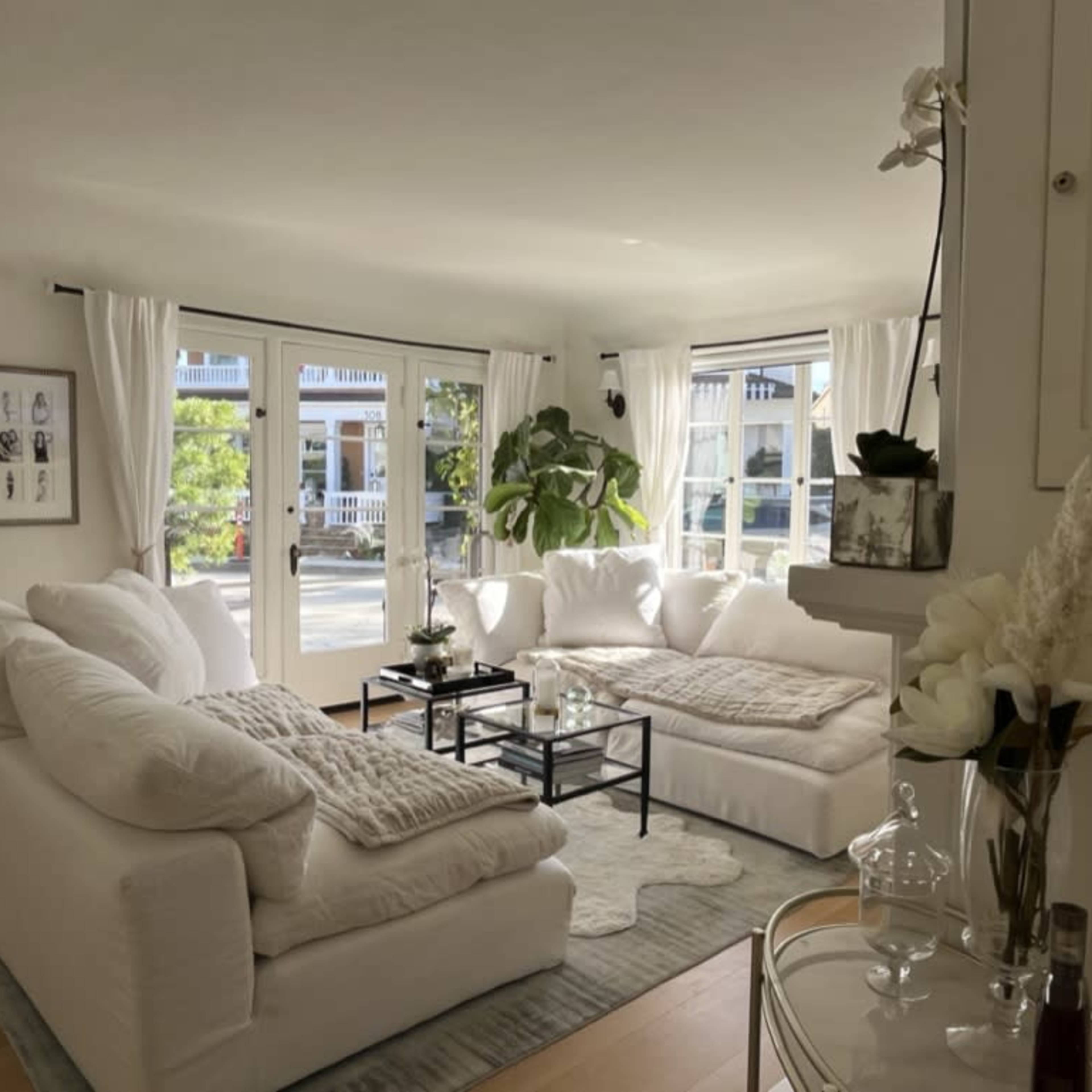 A bright living room features a large white sectional sofa, a coffee table with decorative items, and large windows allowing natural light to enter.
