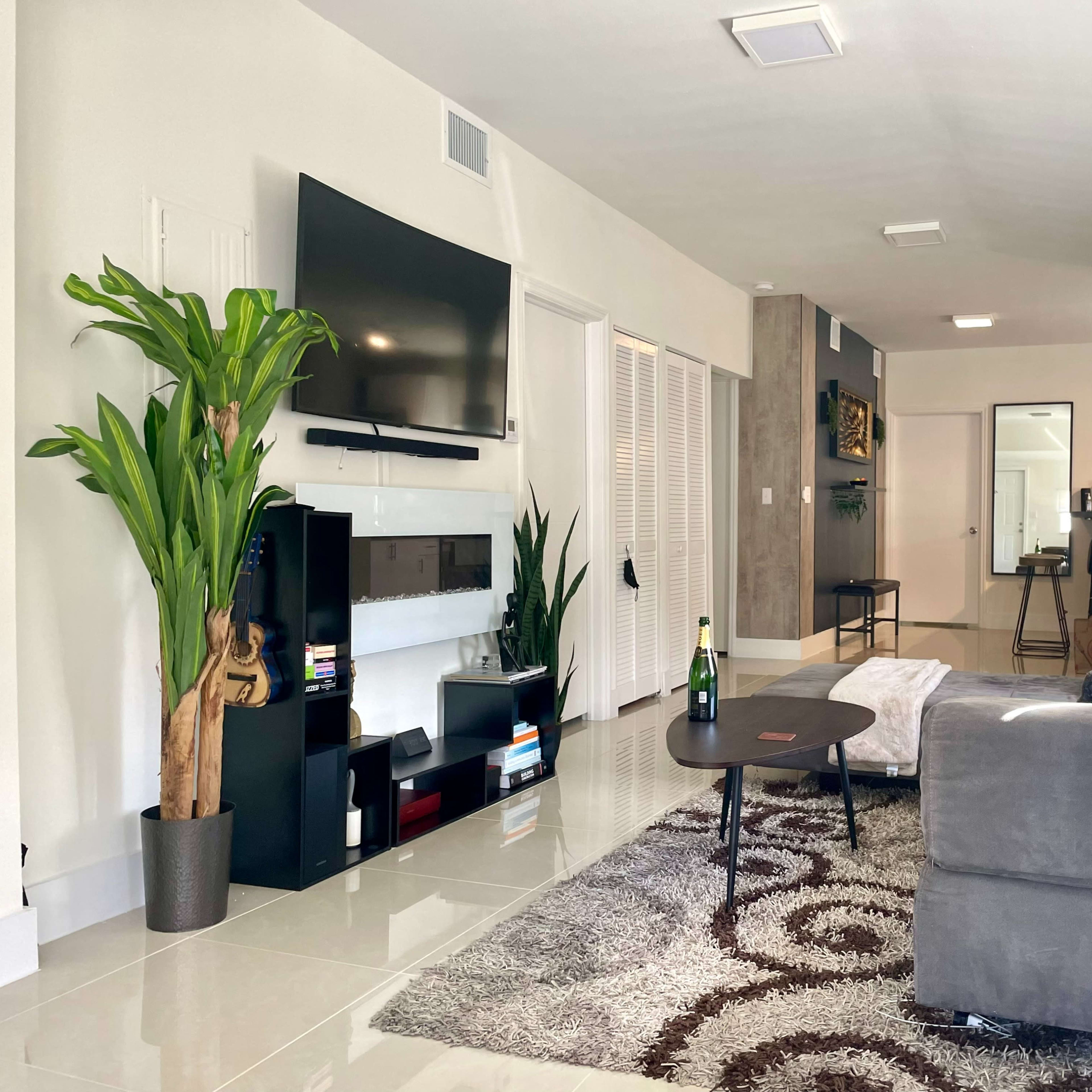 A modern living room with a television mounted on the wall, a gray sofa, and a coffee table on a patterned rug, along with decorative plants and a bar cart.